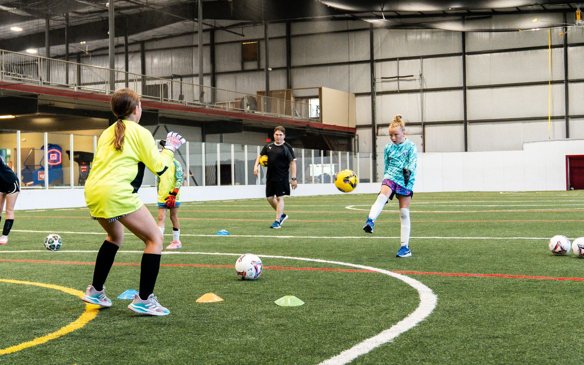 Soccer training: player kicks ball, goalie ready, others observe on indoor field.
