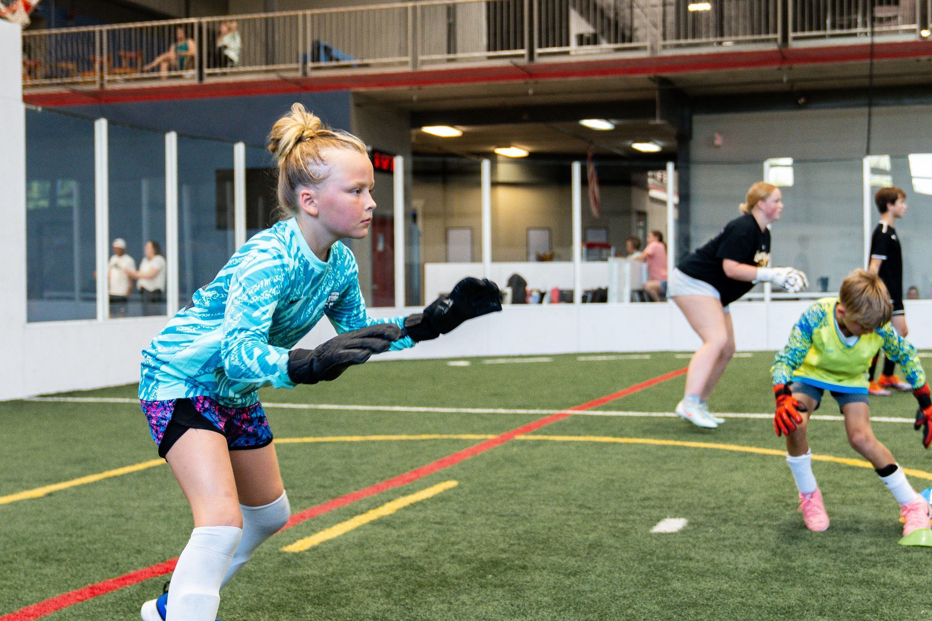 Girl in goalie gear on an indoor soccer field, other players in background.