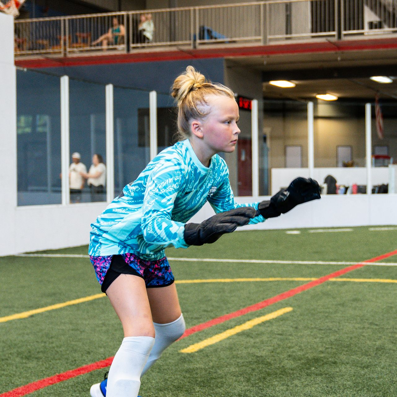 Young soccer goalie in blue uniform, black gloves, on indoor field, focused, ready to catch a ball.