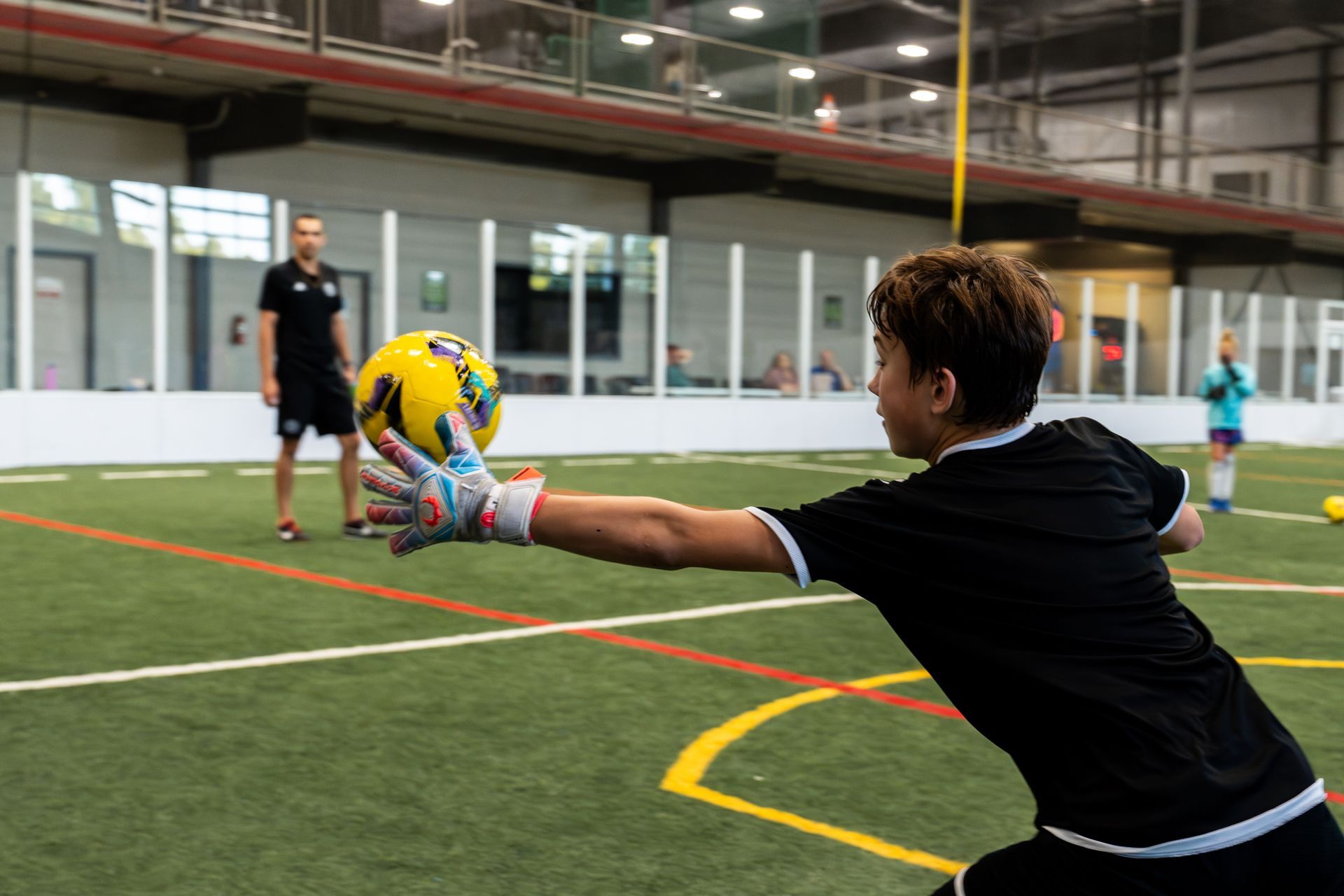 Young soccer goalie in black uniform reaches for a yellow and black ball indoors on a green turf field.