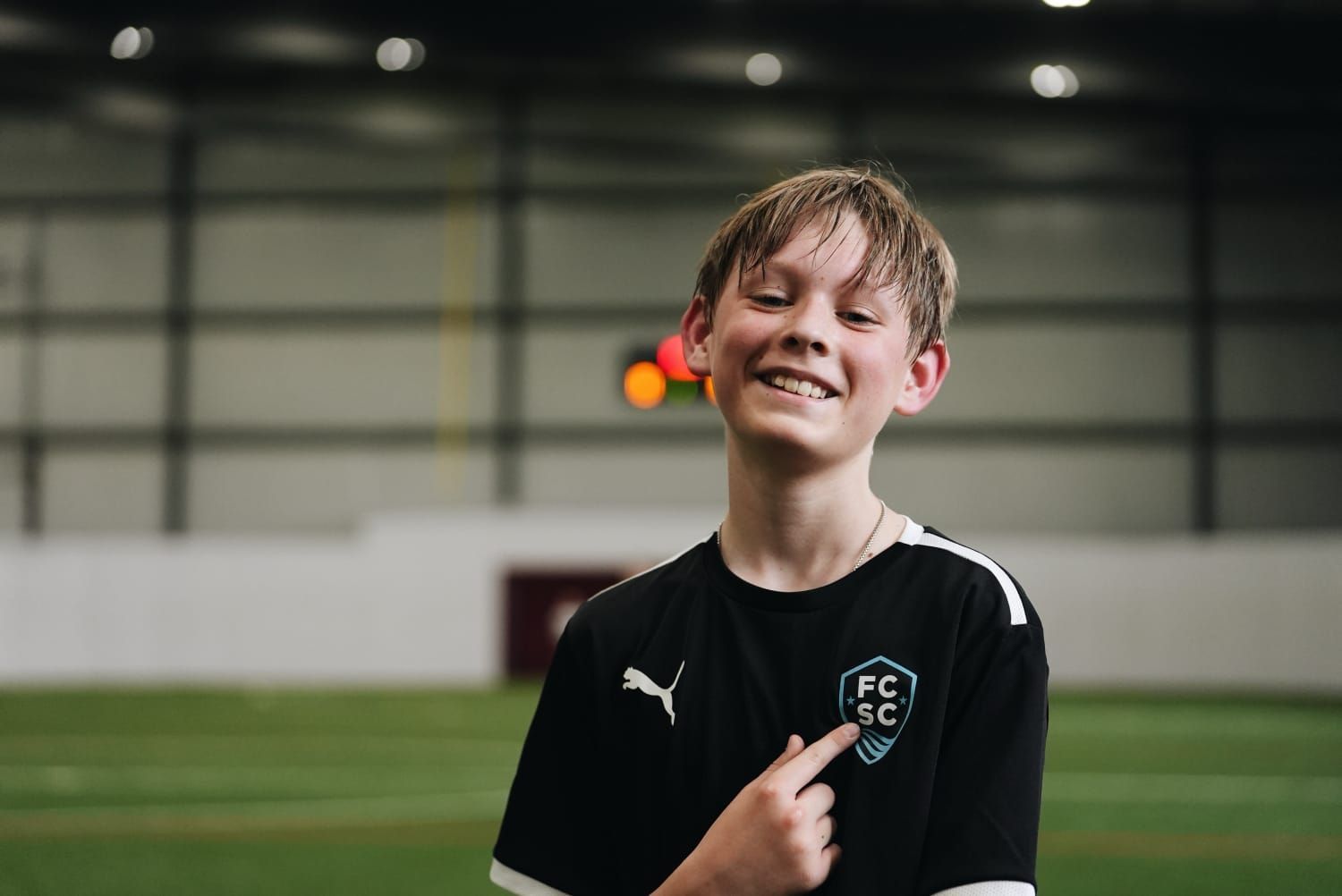 Boy in black soccer jersey points to the team logo with a smile, indoor sports field in the background.