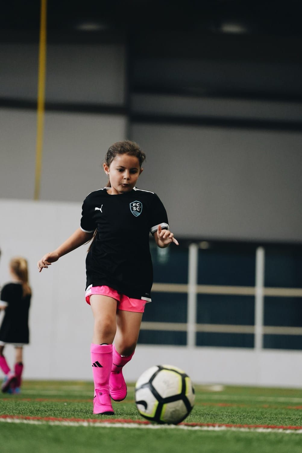 Girl in black jersey and pink shorts dribbles soccer ball indoors on green turf.