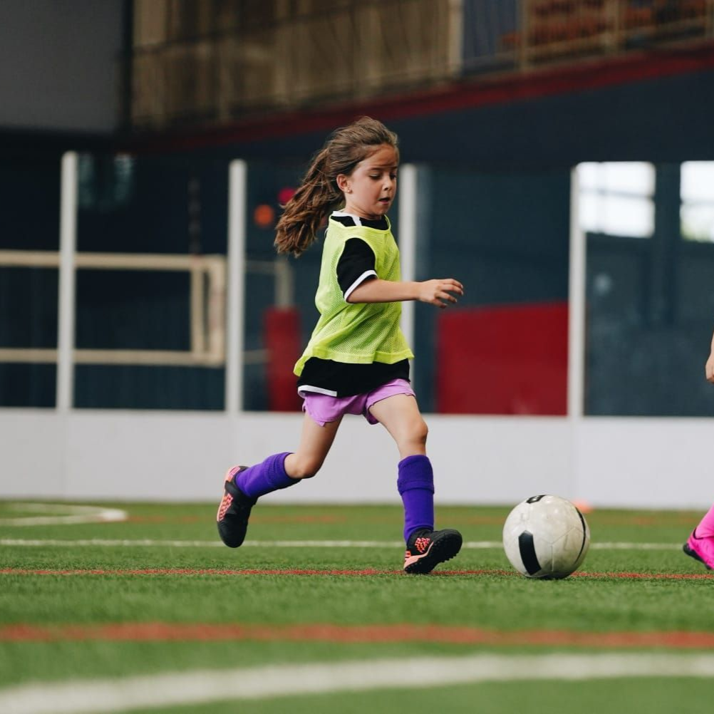 Girl in green shirt kicks soccer ball on indoor field.