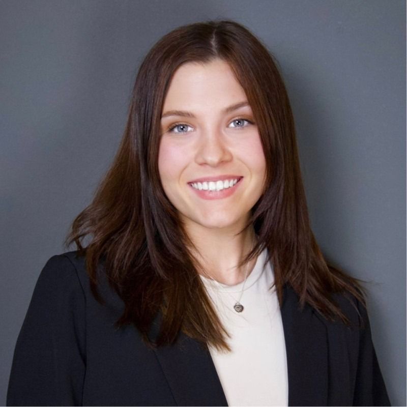 Woman with brown hair, smiling, wearing a black blazer and white top, against a gray background.