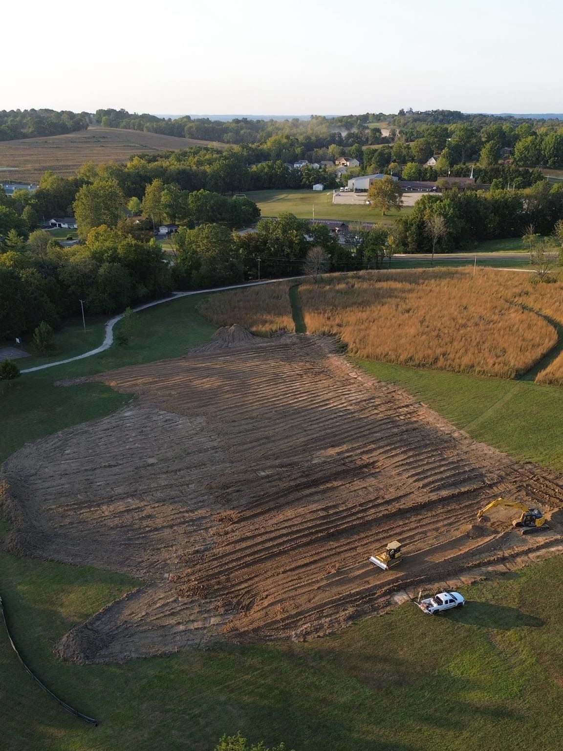 Field being worked on by construction equipment, next to green trees and a path.