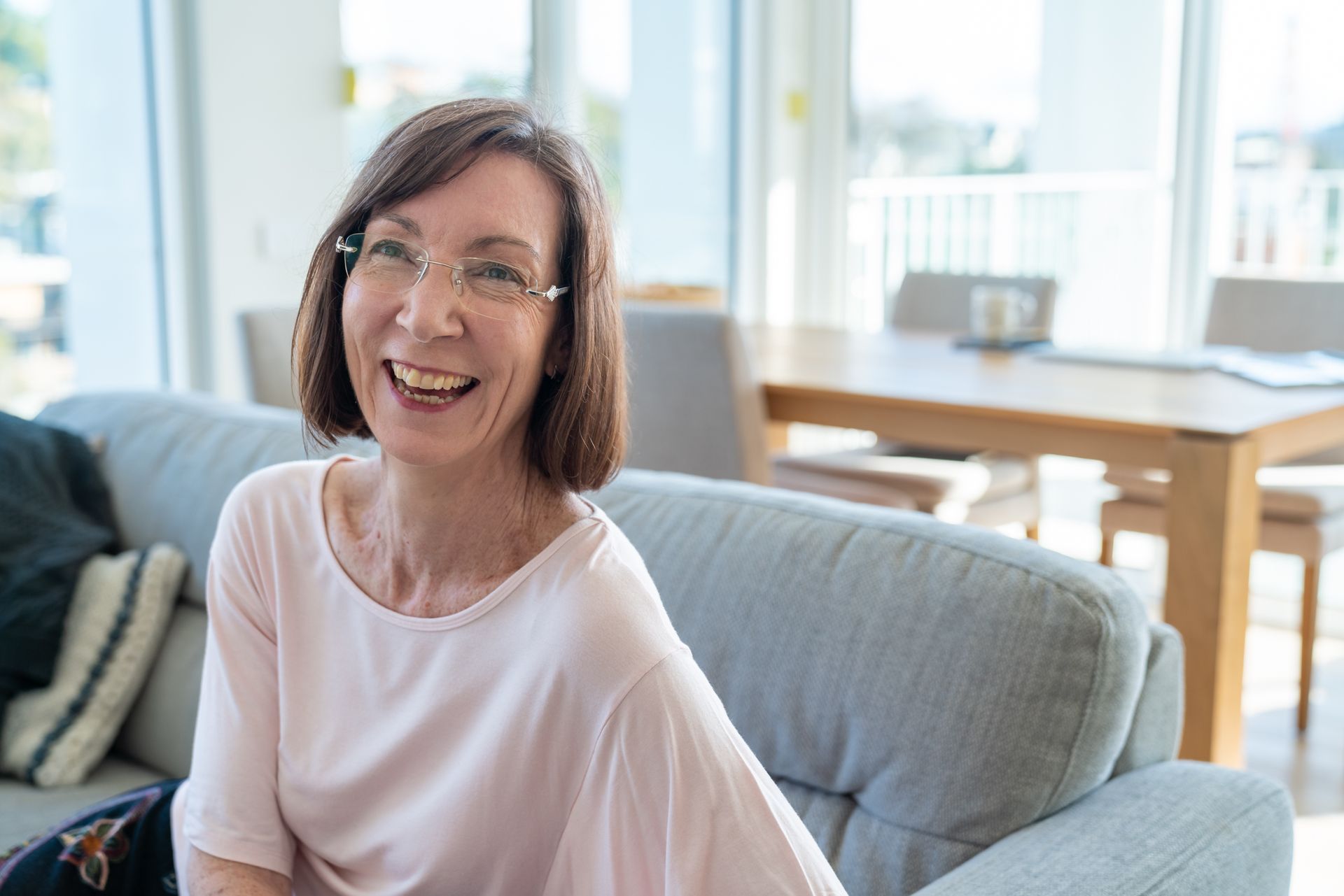 Woman smiling, sitting on a gray couch in a bright living room. She wears glasses and a pink shirt.