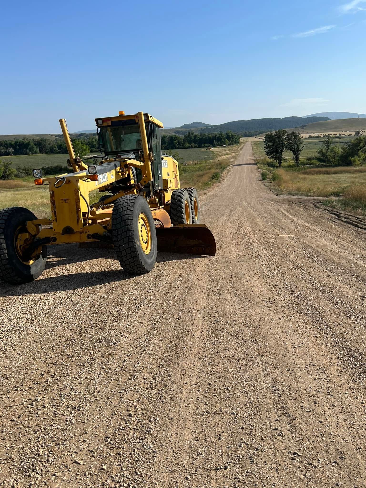 Yellow road grader on a gravel country road under a clear blue sky