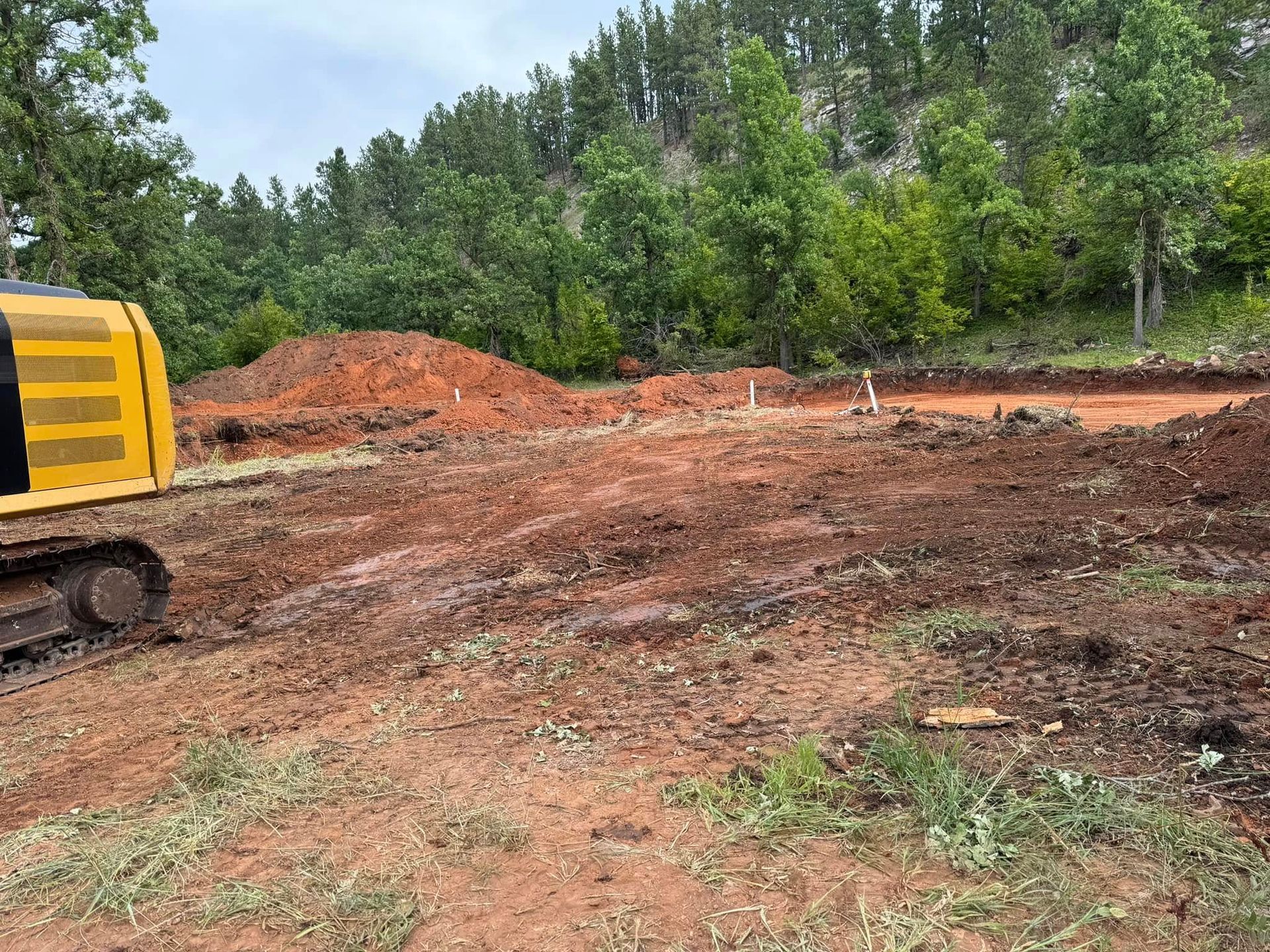 Construction site with yellow excavator on muddy red dirt, backed by trees and a wooded hillside