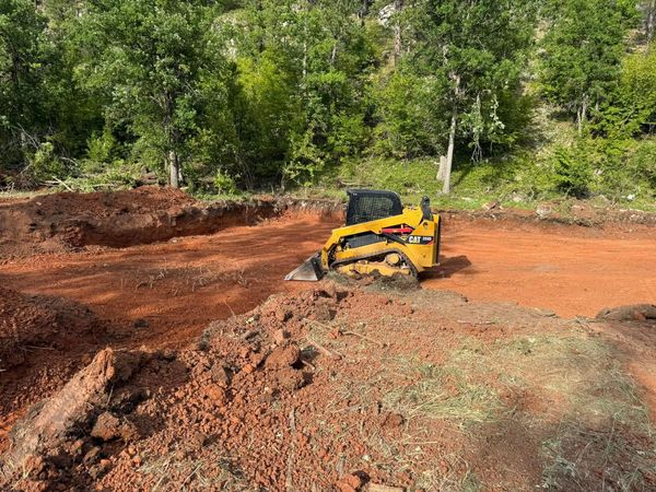 Yellow skid steer clearing red dirt in a wooded clearing.