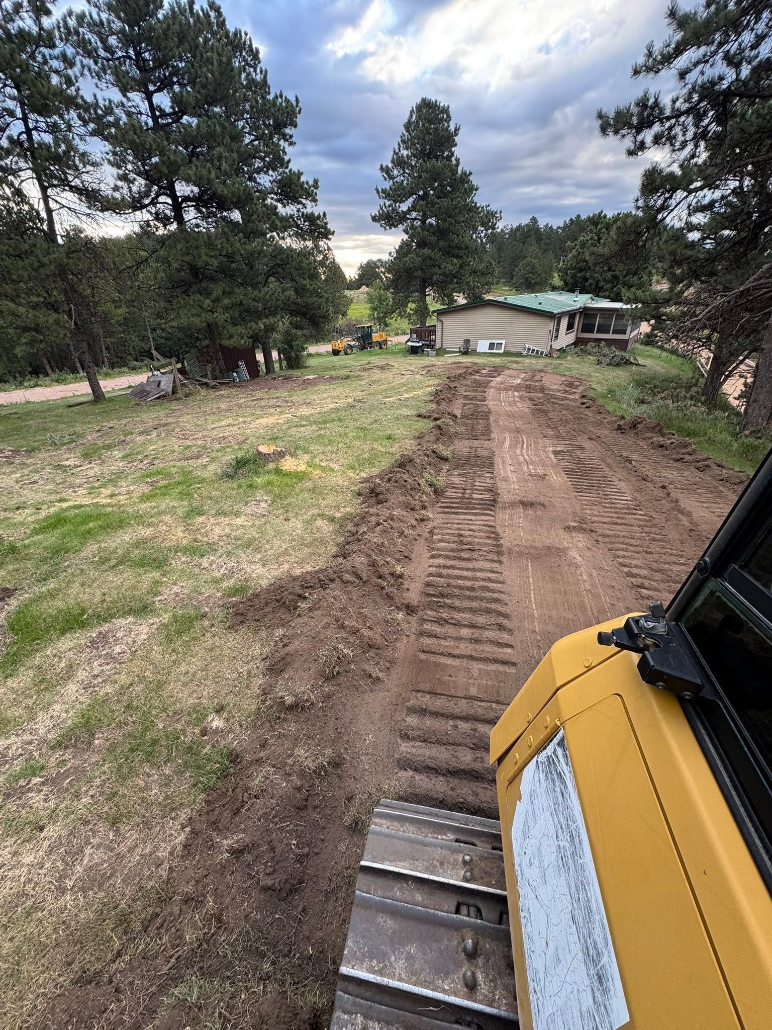 Dirt road being graded beside trees, viewed from a yellow construction vehicle under a cloudy sky