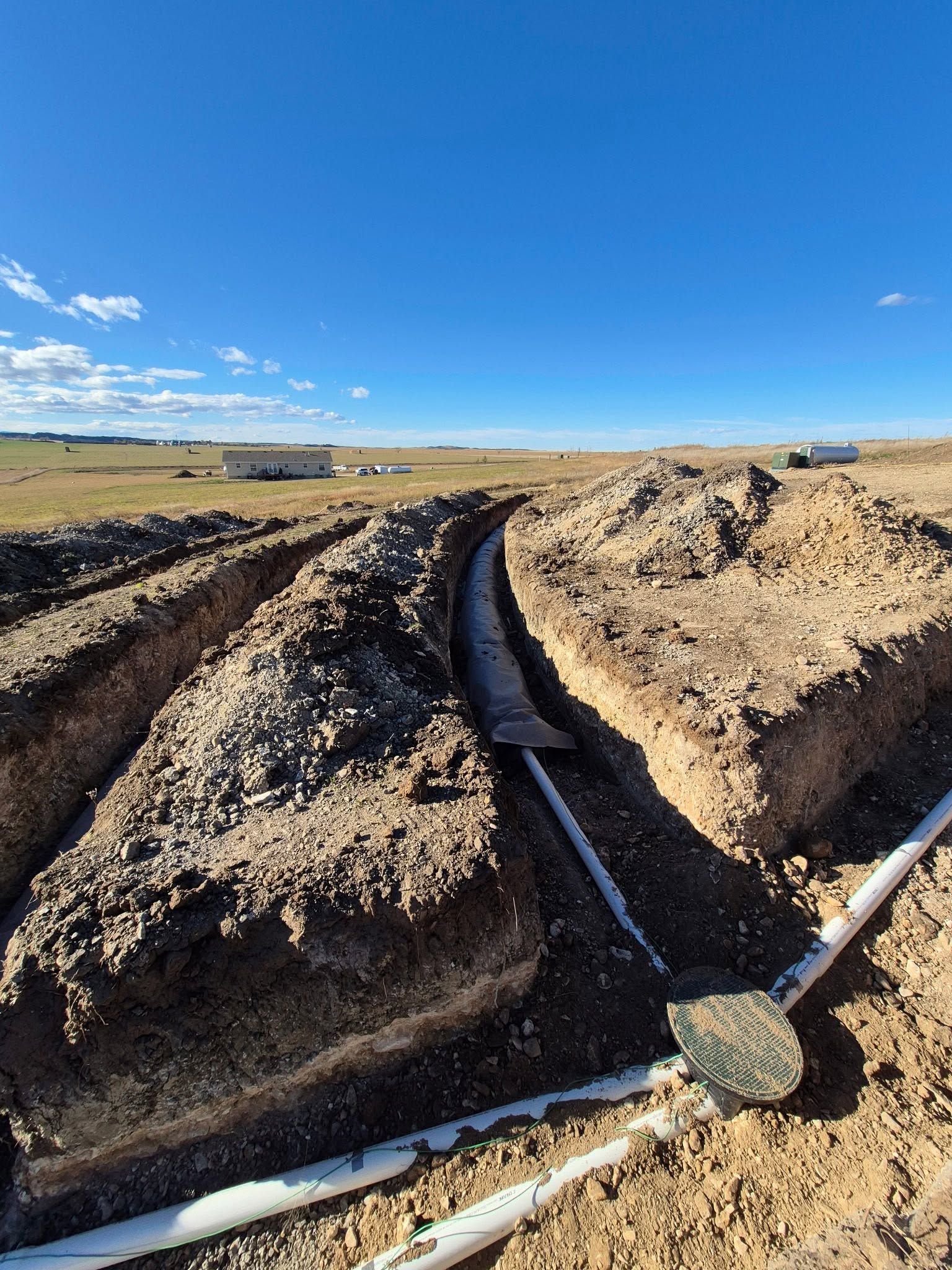 Ditch with black drainage pipe running through muddy earth under a clear blue sky in a rural field