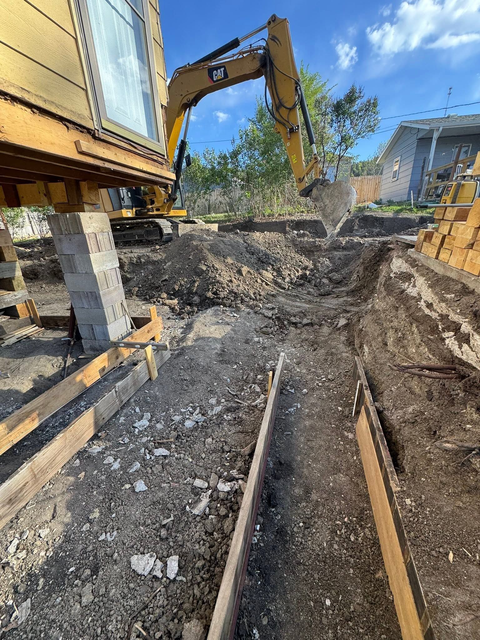 Construction site with excavator digging a trench beside a wooden house foundation