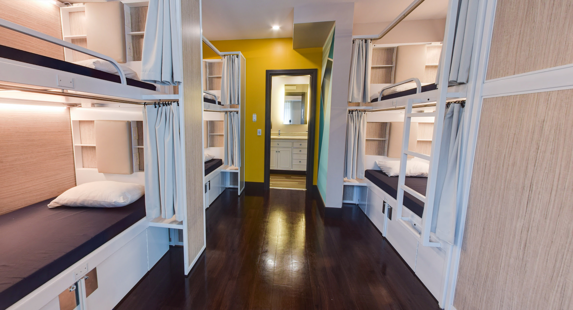 Chambre de dortoir avec lits superposés ; parquet foncé, mur jaune et salle de bains au loin.