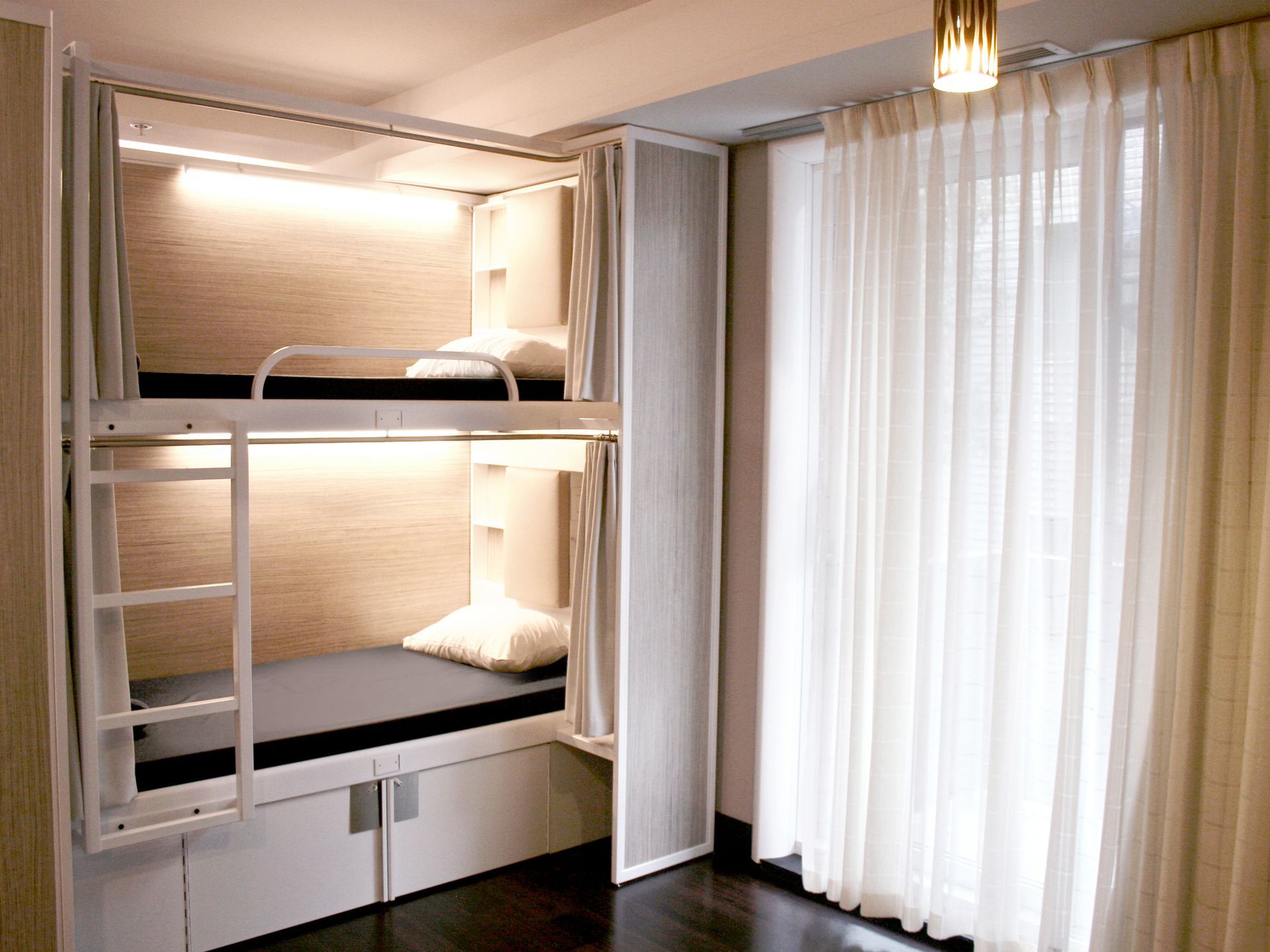 Lits superposés dans une chambre moderne de style capsule avec rideaux blancs et éclairage chaleureux.