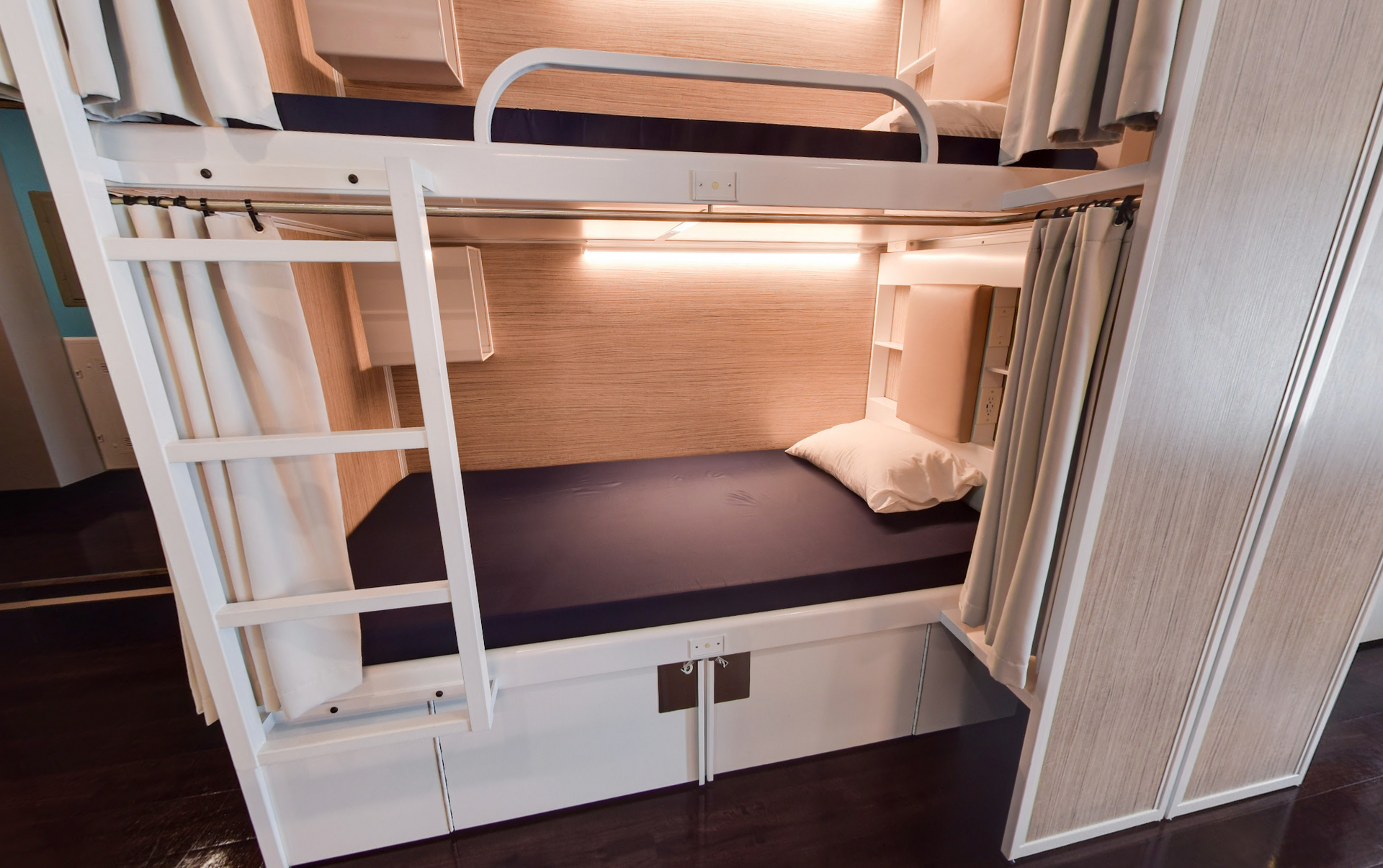 Lits superposés dans une pièce lumineuse avec une échelle, cadre blanc, matelas bleus et rangements en dessous.