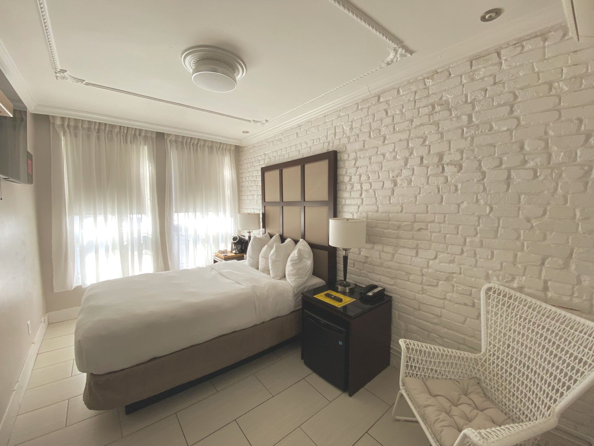Chambre avec mur en briques blanches, lit avec tête de lit, chaise et fenêtres avec voilages.