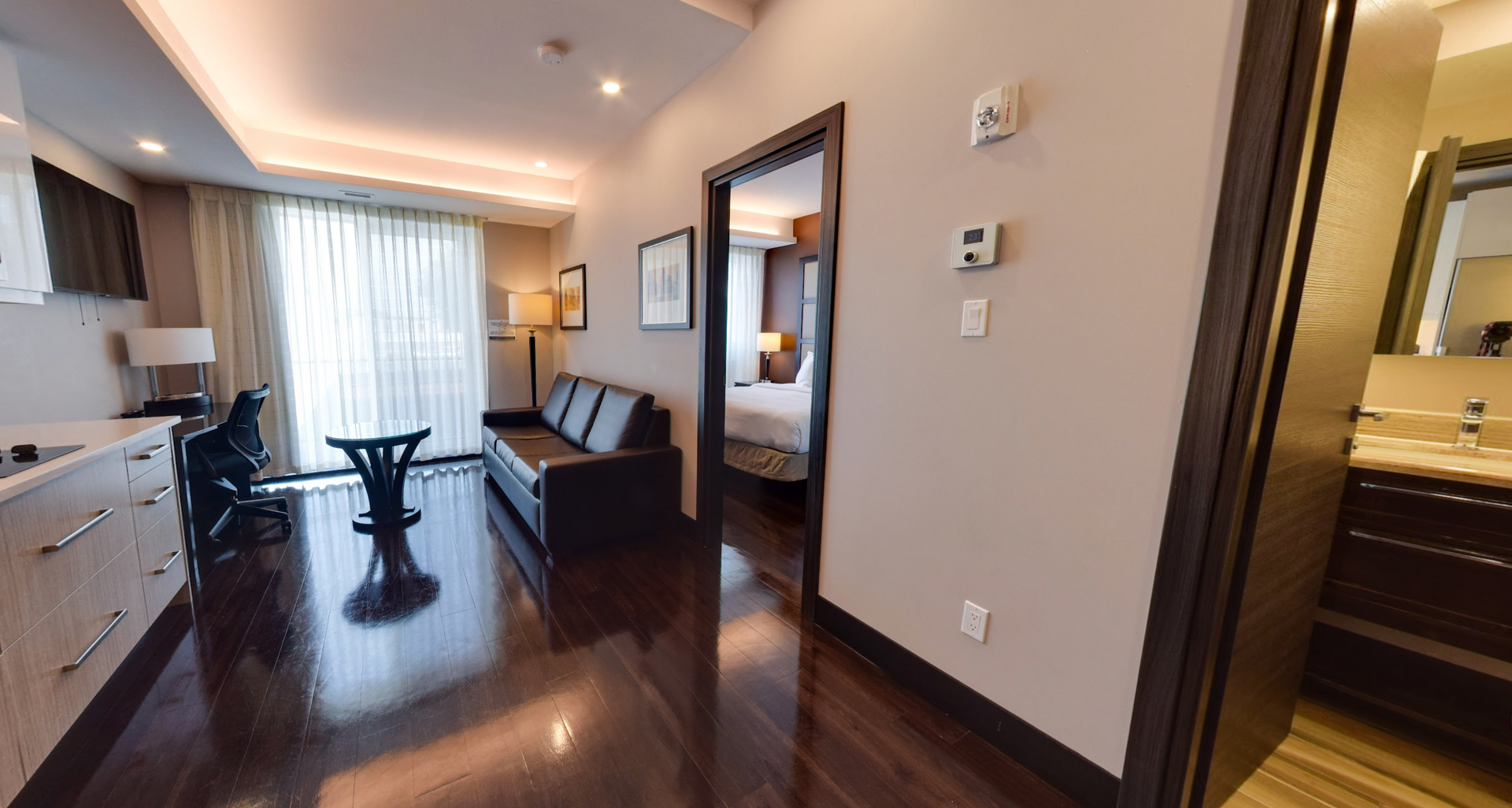 Suite d'hôtel spacieuse : coin salon avec canapé, bureau et porte donnant sur la chambre et la salle de bains. Parquet foncé.