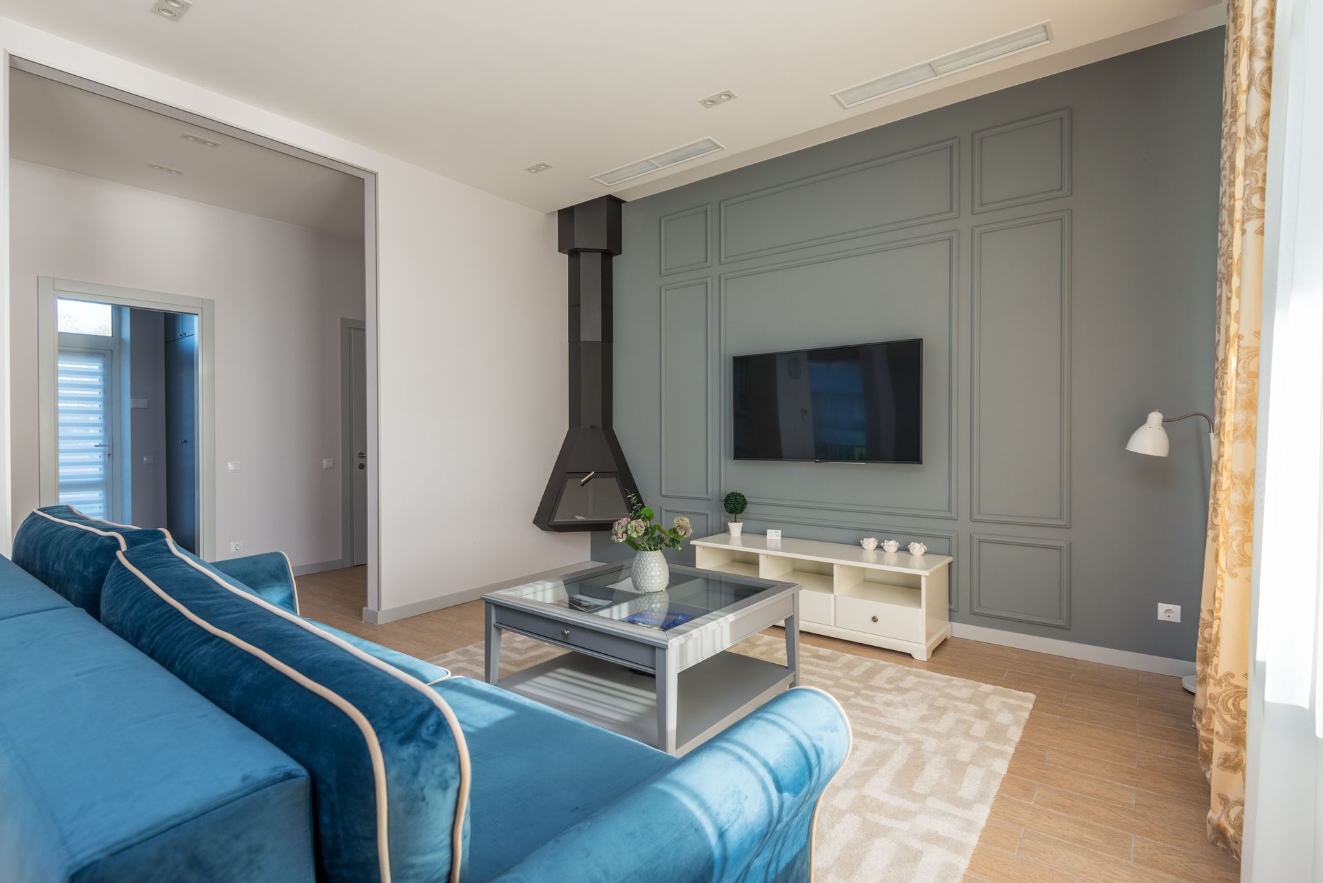 Modern living room with blue sofa, gray paneled wall, wall-mounted TV, and black fireplace.