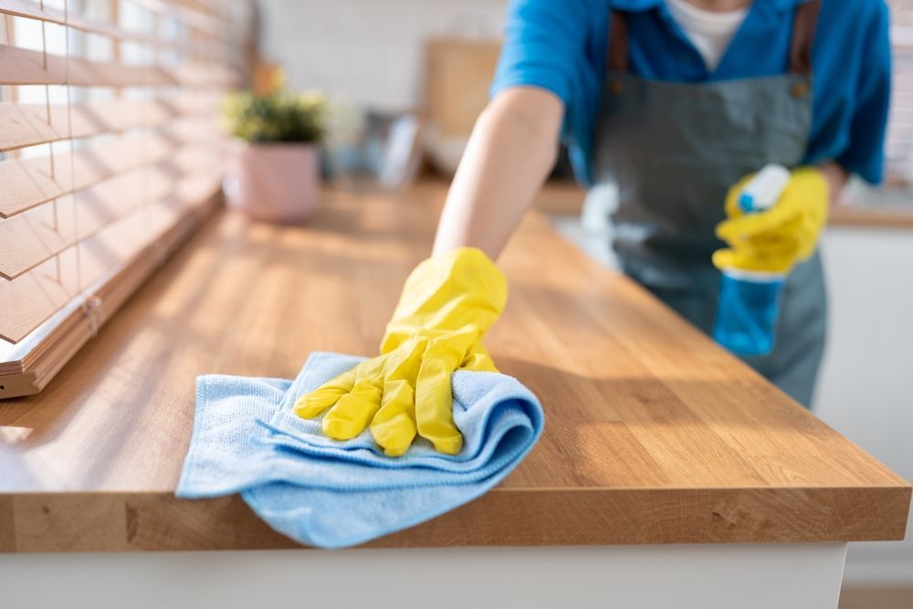 Person wiping a wooden countertop with a blue cloth and yellow gloves in a bright kitchen