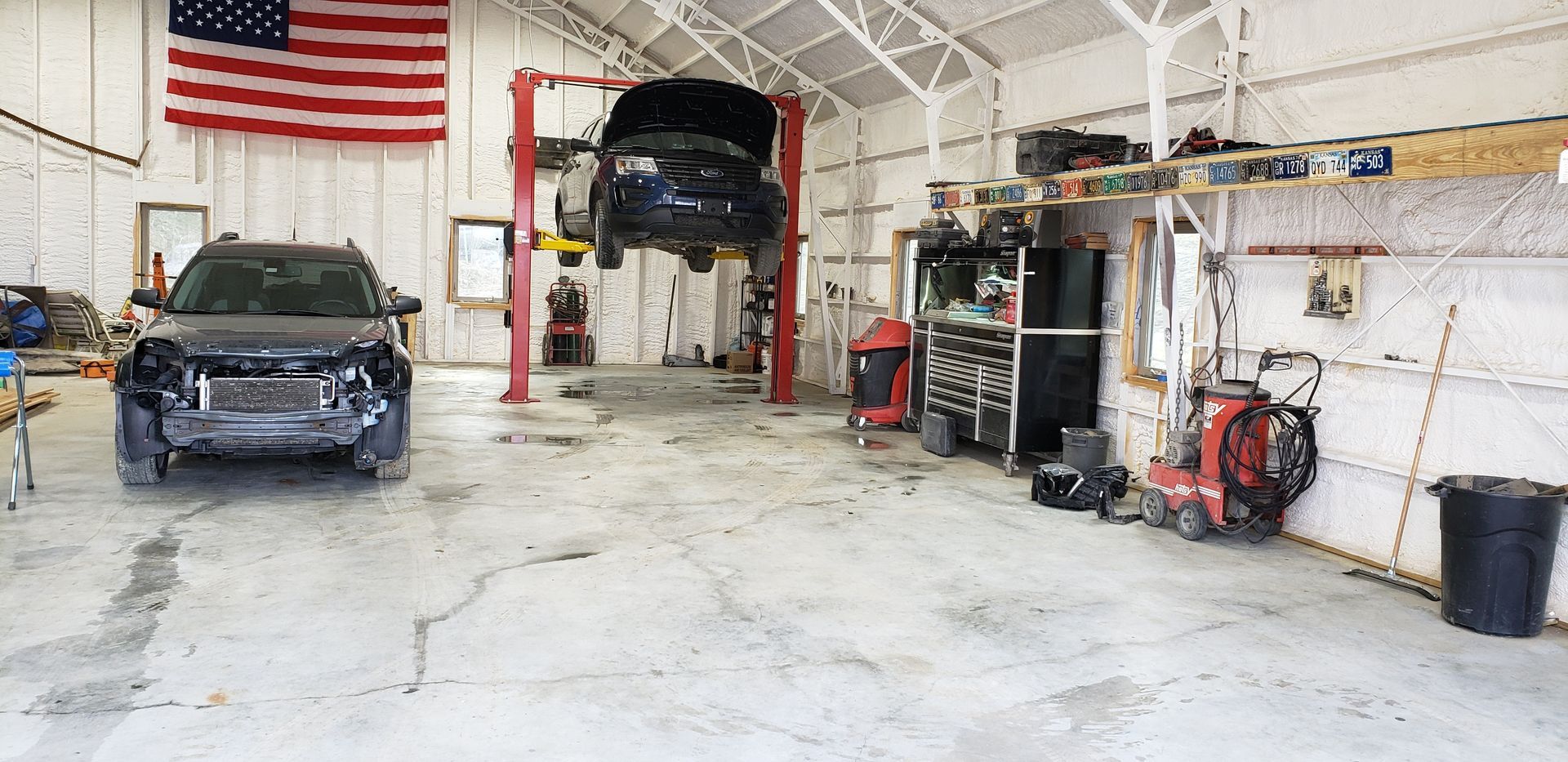 Auto shop interior with two vehicles, a lift, tools, and a large American flag on the wall