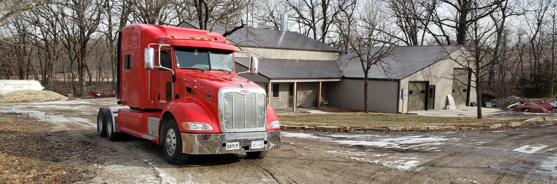 Red semi-truck parked on a muddy lot in front of a house and garage on a cloudy day