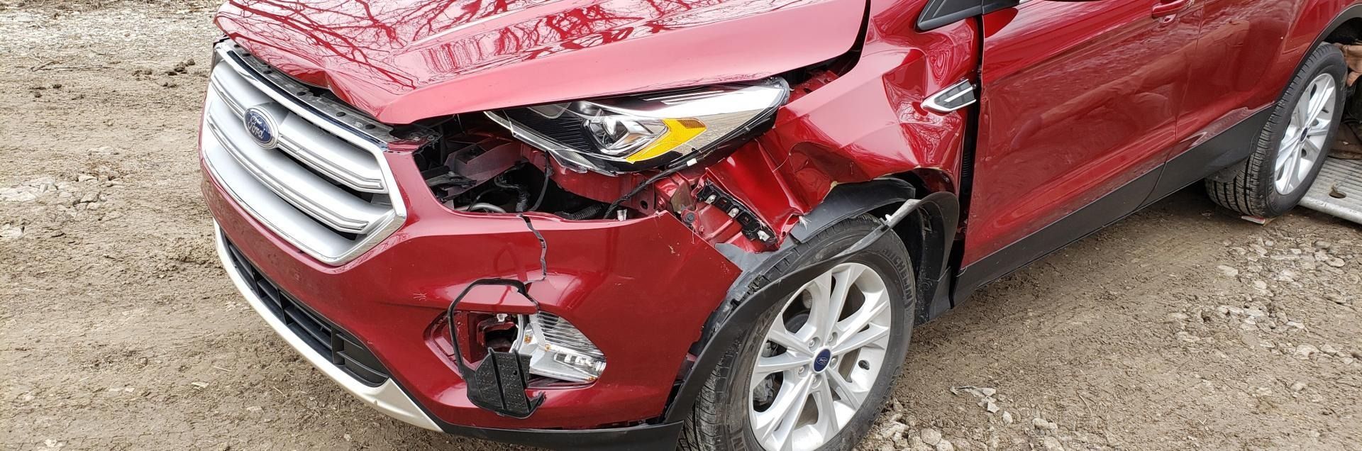 Red car with severe front-left body damage, crumpled bumper and broken headlight on a dirt surface.