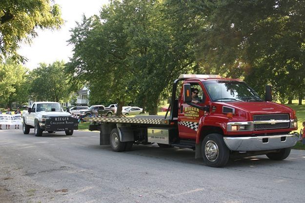 Red flatbed tow truck parked on a tree-lined street with a white pickup behind it.
