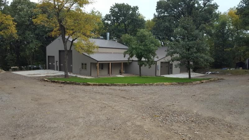 Gray house with porch beside a gravel lot, surrounded by trees under an overcast sky