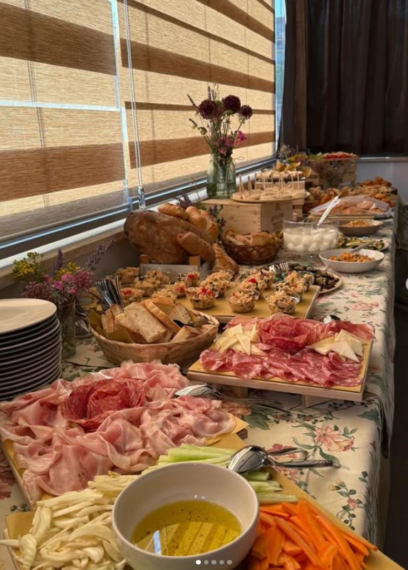 Tavolo da buffet con salumi, formaggi, pane, verdure, salse e una composizione floreale.