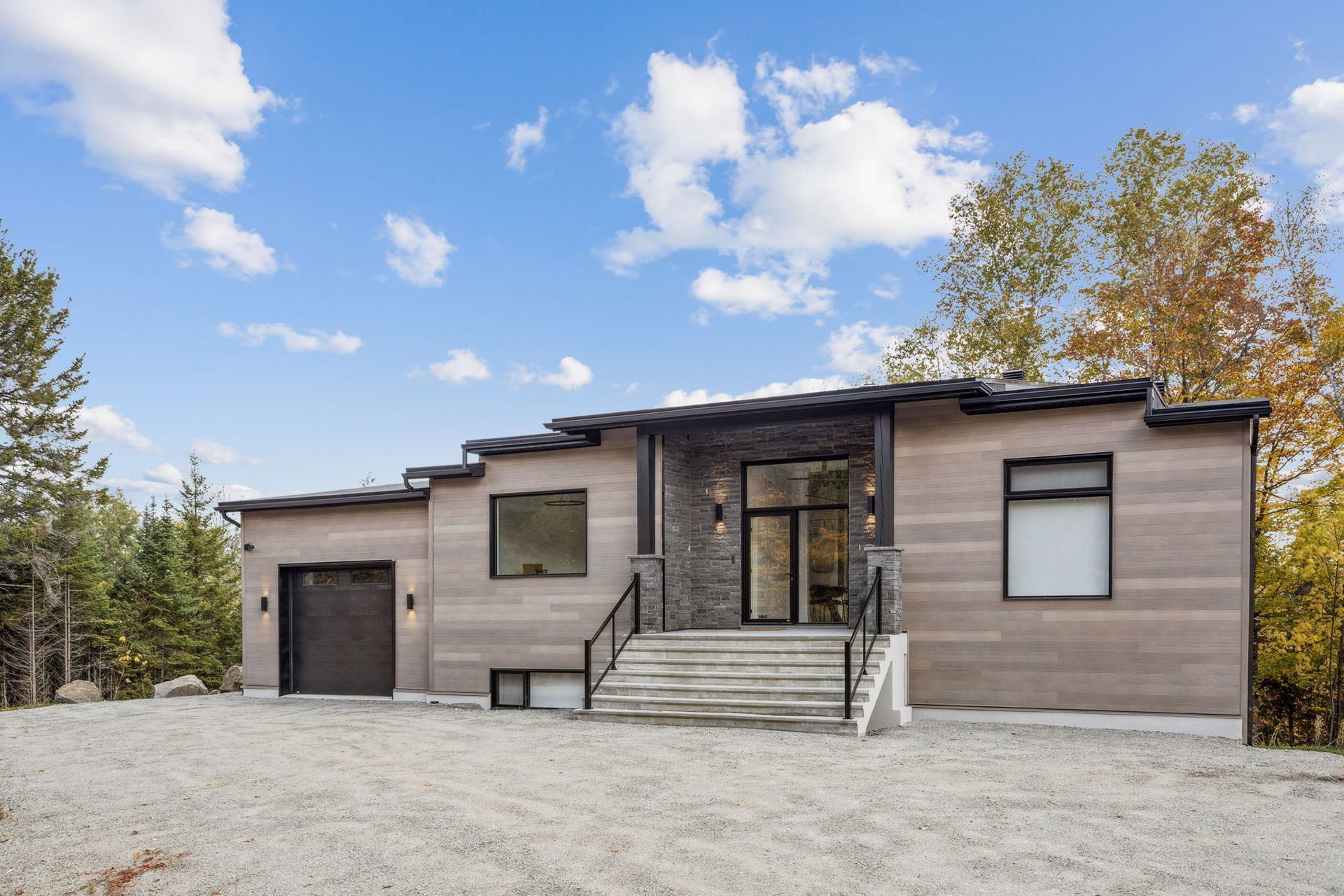 Maison moderne avec un revêtement extérieur brun clair, des éléments en pierre, des finitions noires et une allée en gravier gris.