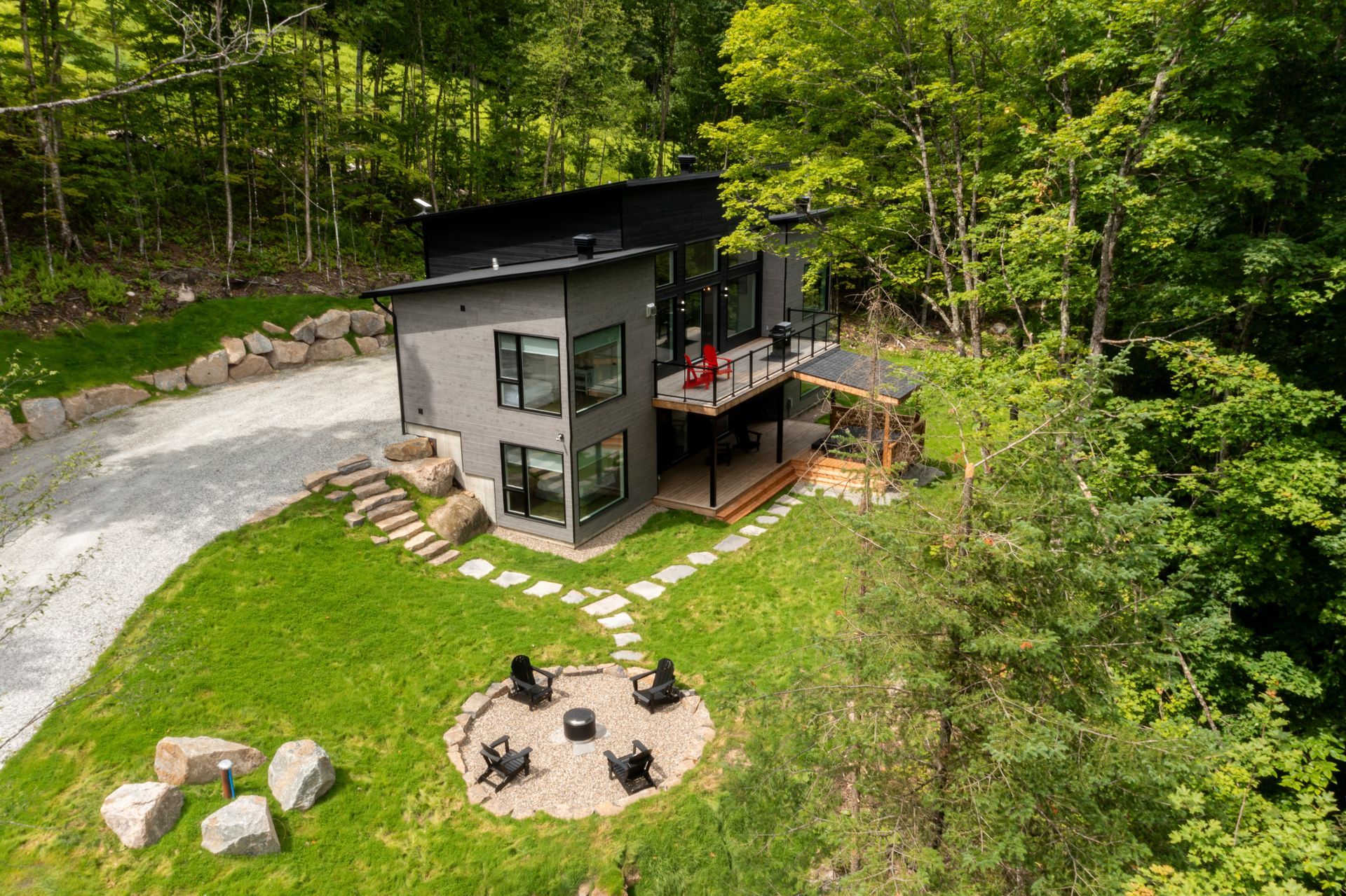 Maison grise moderne nichée dans une forêt, avec terrasse, foyer extérieur et allée de gravier.