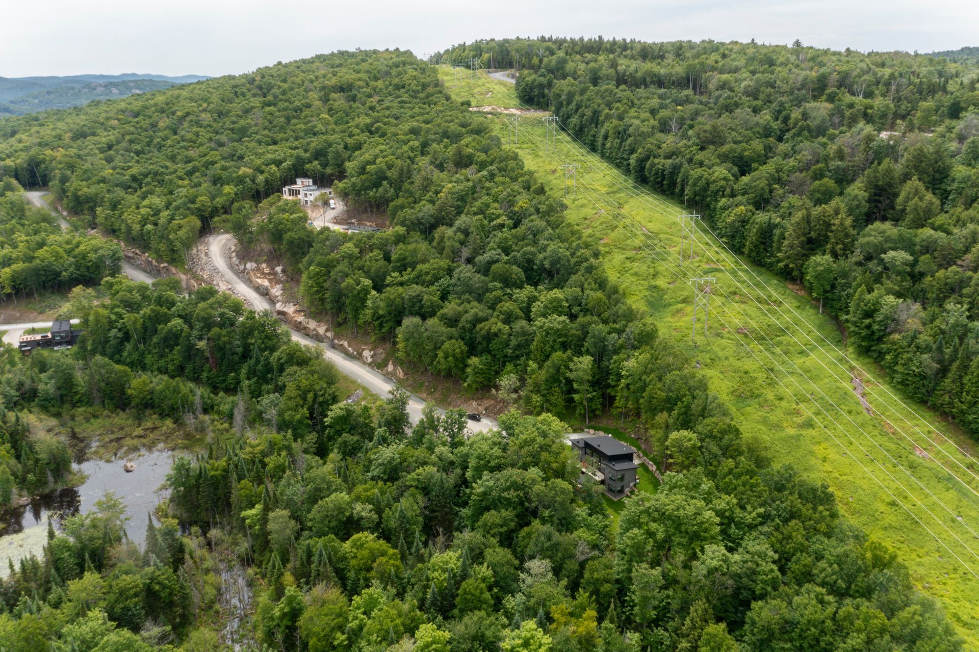 Colline verdoyante traversée par un large chemin dégagé ; une route serpente à proximité. La forêt entoure le chemin, une petite construction est visible.