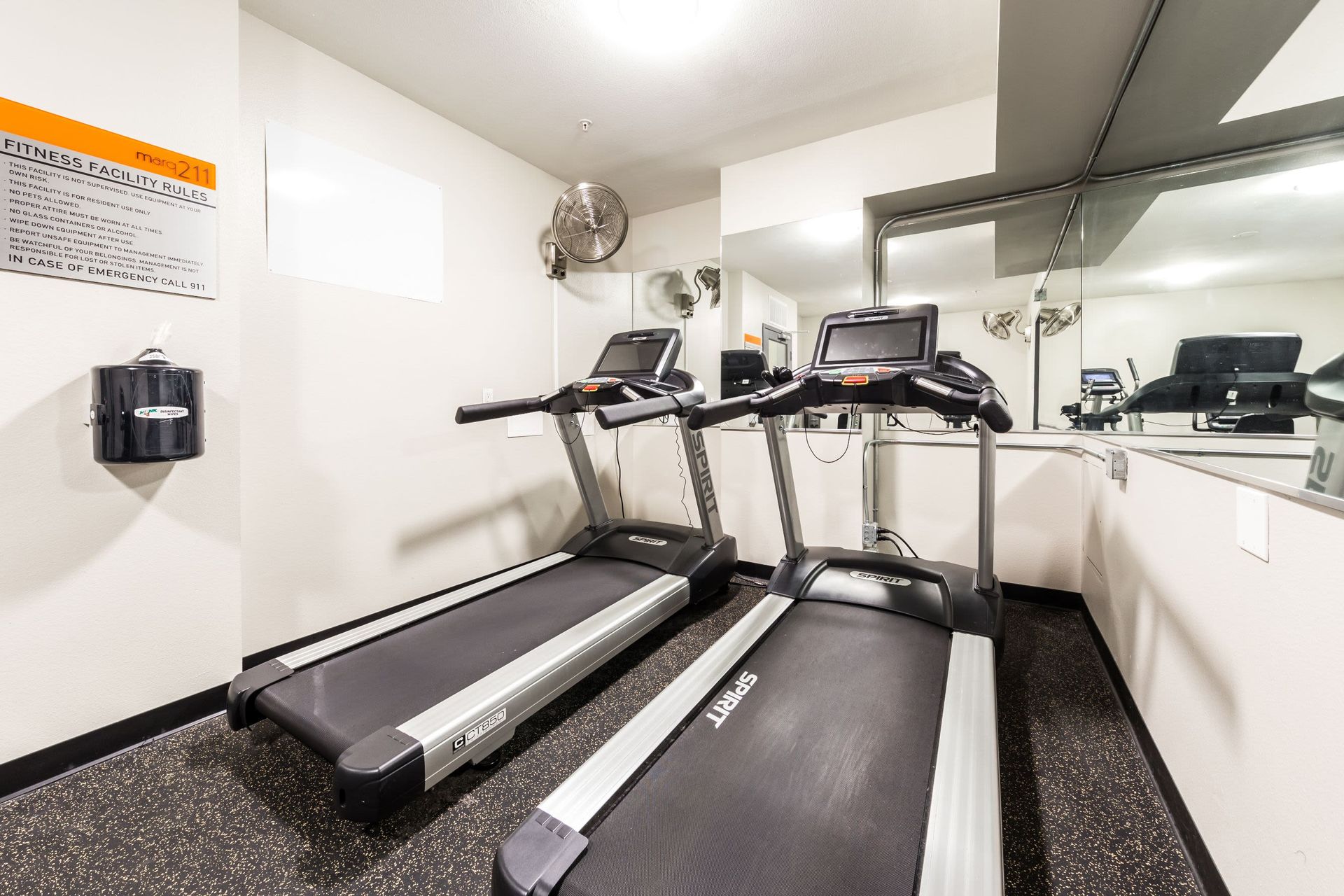 Interior view of a compact fitness facility with two treadmills in the foreground and a stationary exercise bike in the background. A wall-mounted fan, sanitizer dispenser, and posted fitness facility rules are visible on the left. The room has large mirrors reflecting the exercise equipment, enhancing the sense of space at Marq 211 in Seattle, WA.