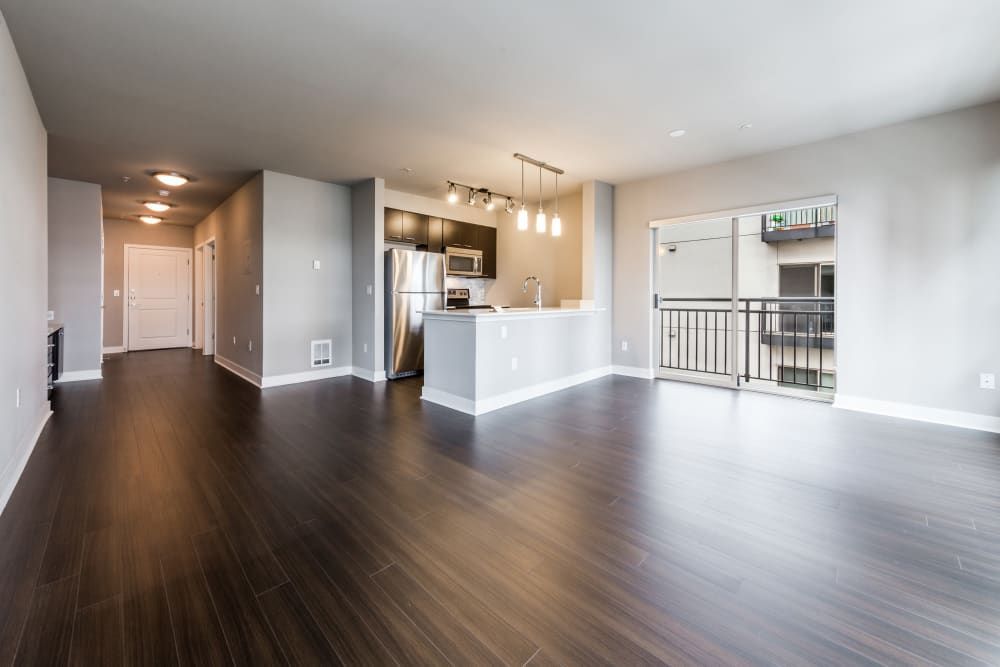 Spacious and modern open-plan apartment interior featuring dark hardwood floors, a kitchen area with stainless steel appliances and a white countertop island, a bright living space with recessed lighting, and sliding glass doors leading to a balcony at Marq 211 in Seattle, WA.