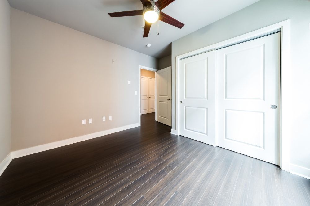 An empty room featuring glossy dark wood flooring, a pale beige wall with electrical outlets, a ceiling fan overhead, and a large two-panel white closet door on the right. Another open door is visible in the background leading to a room with a lit ceiling light at Marq 211 in Seattle, WA.