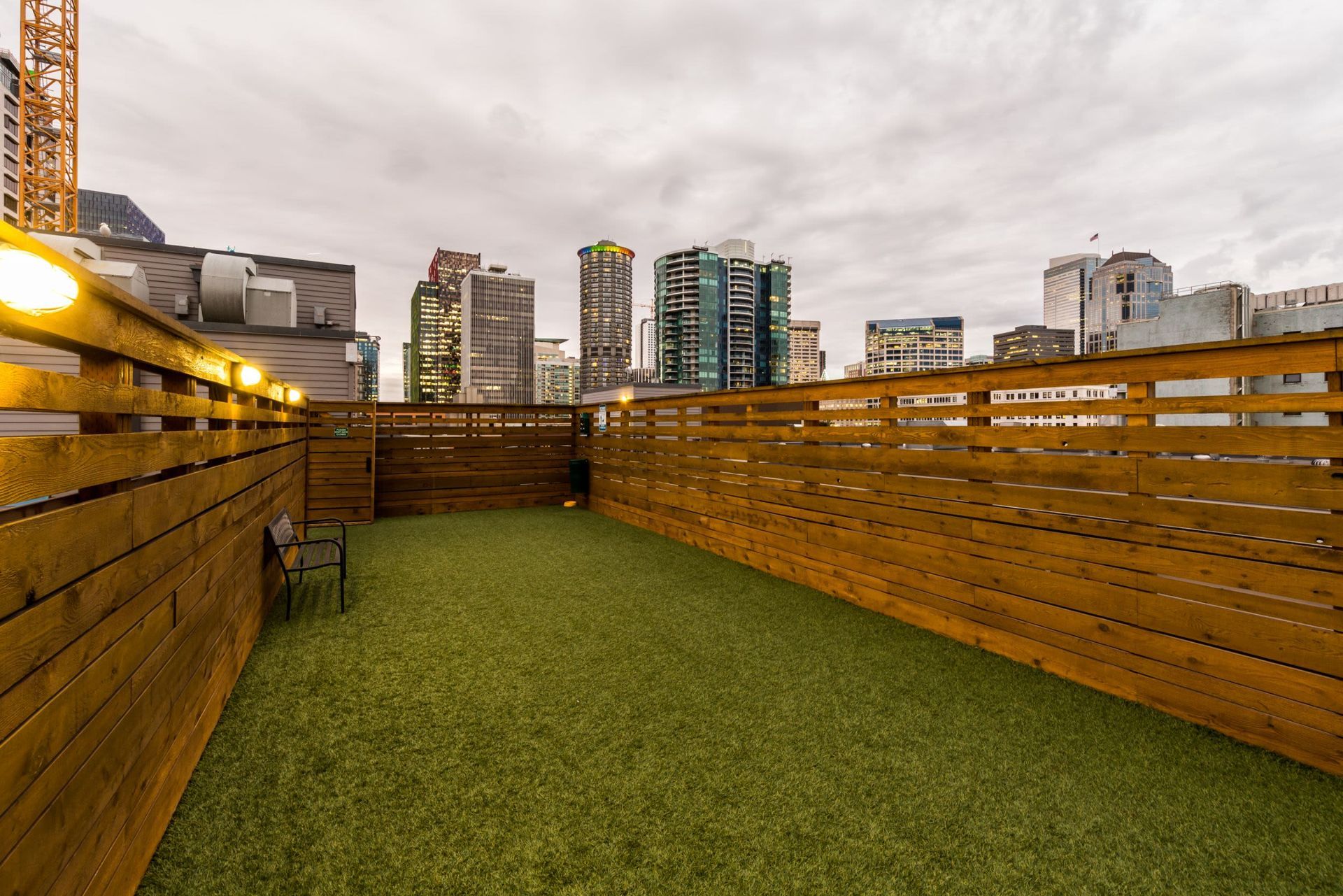 Rooftop dog park with artificial grass and wooden fencing, adorned with string lights, offering a view of a bustling city skyline with various modern high-rise buildings, under a cloudy sky at Marq 211 in Seattle, WA.
