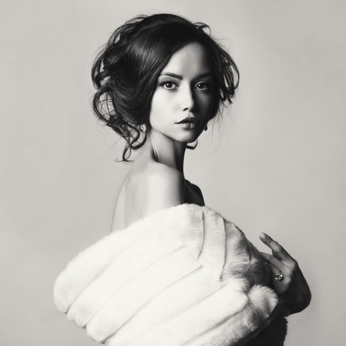 Ritratto in bianco e nero di una persona con un'elegante acconciatura raccolta, avvolta in una stola di pelliccia bianca, che guarda verso la macchina fotografica.