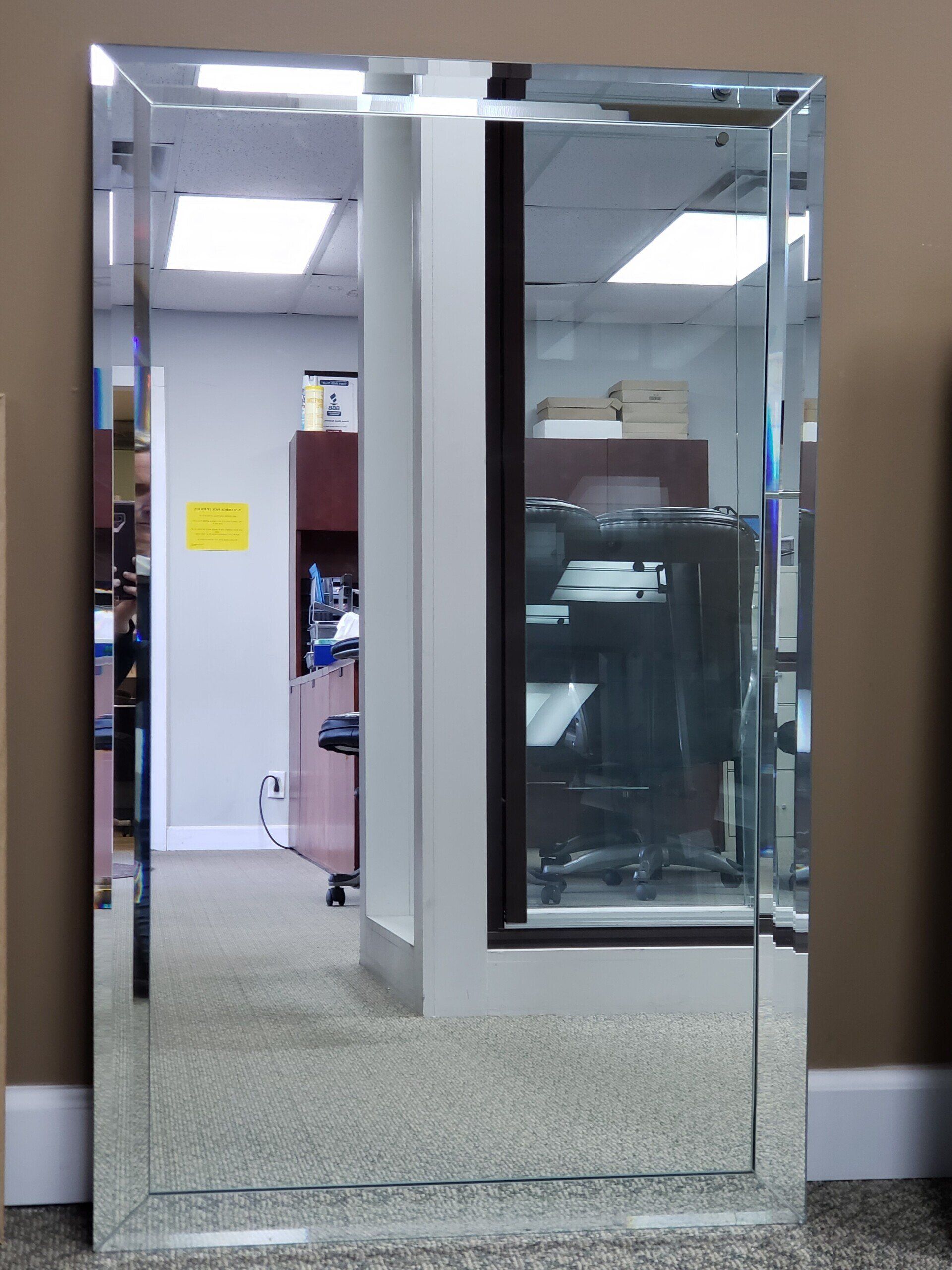 Full-length rectangular mirror with beveled edges; reflects an office space with desks and overhead lights.