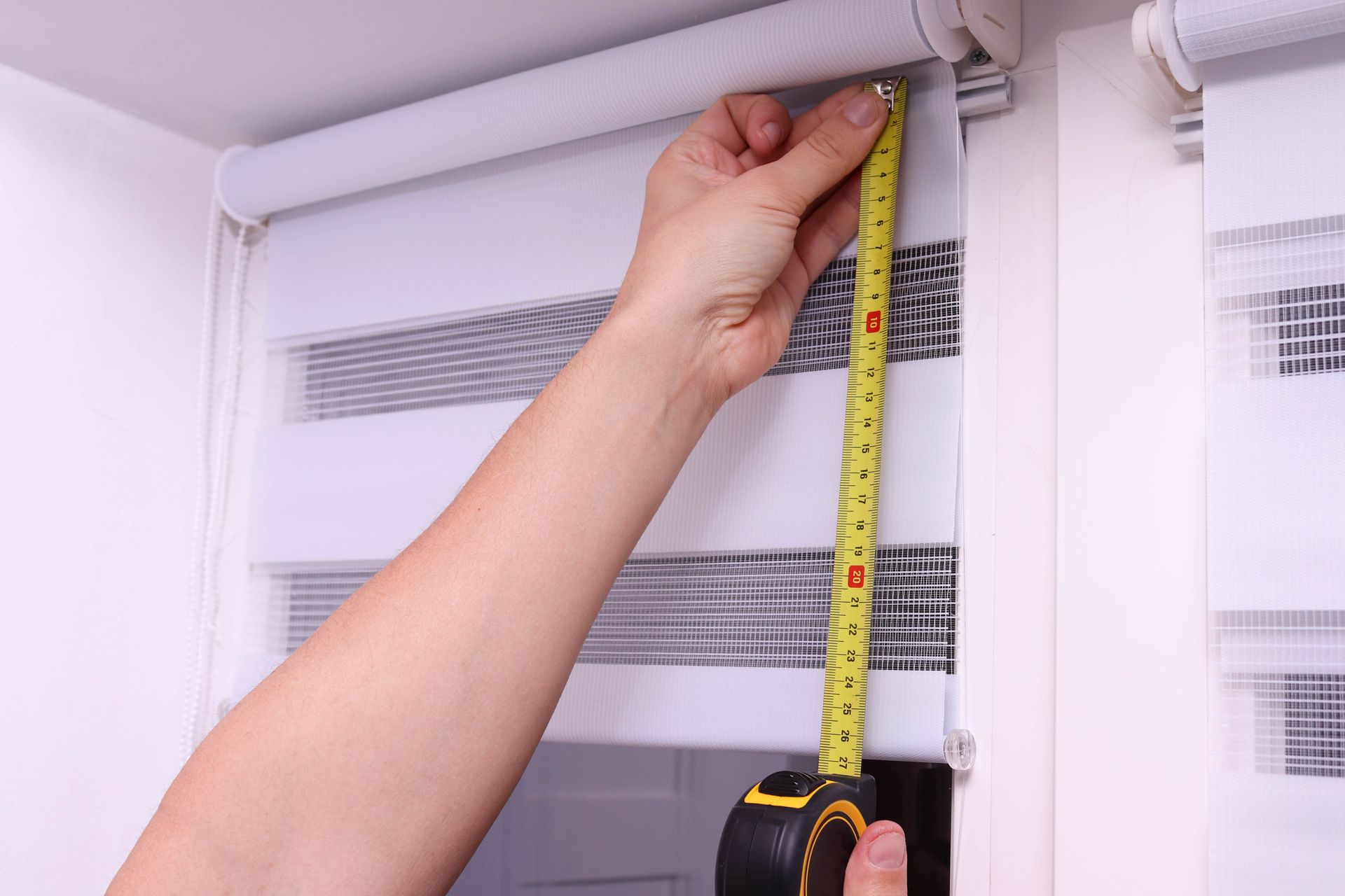 A homeowner measuring the height of white roller blinds on a window with a yellow tape measure