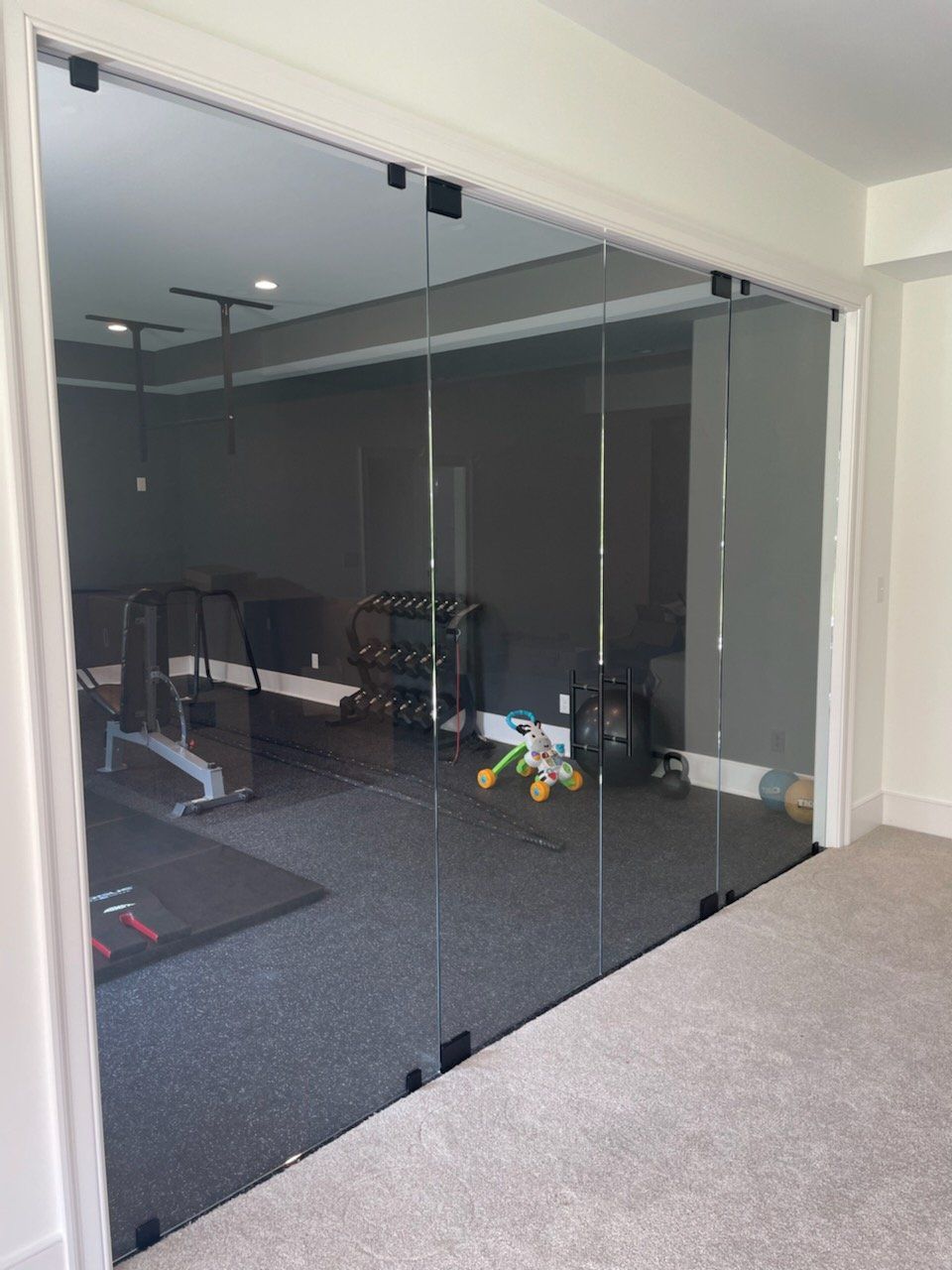 Glass-walled home gym with weights, equipment, and a kid's toy inside; viewed through doorway, gray walls, carpet.
