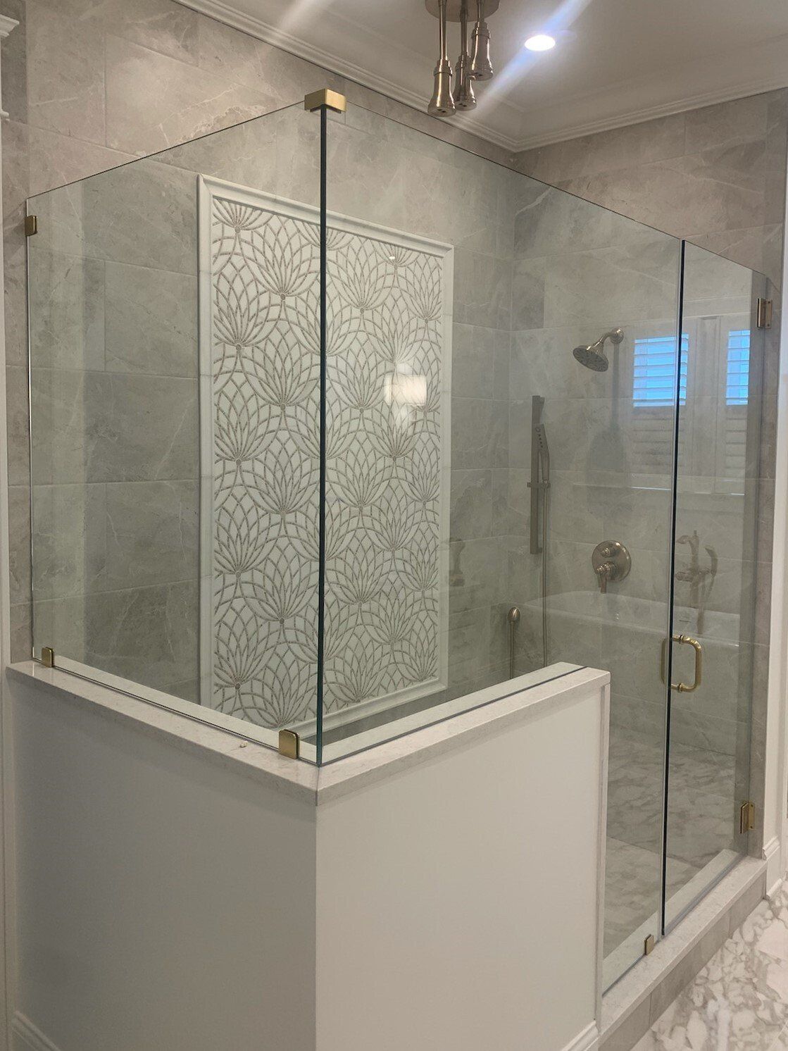 Glass-enclosed shower with gold hardware, white walls, and a decorative tile feature.