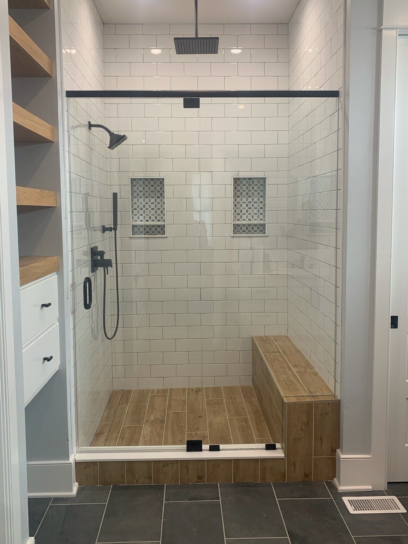 Modern shower with glass door, subway tile, wooden bench, and black fixtures.