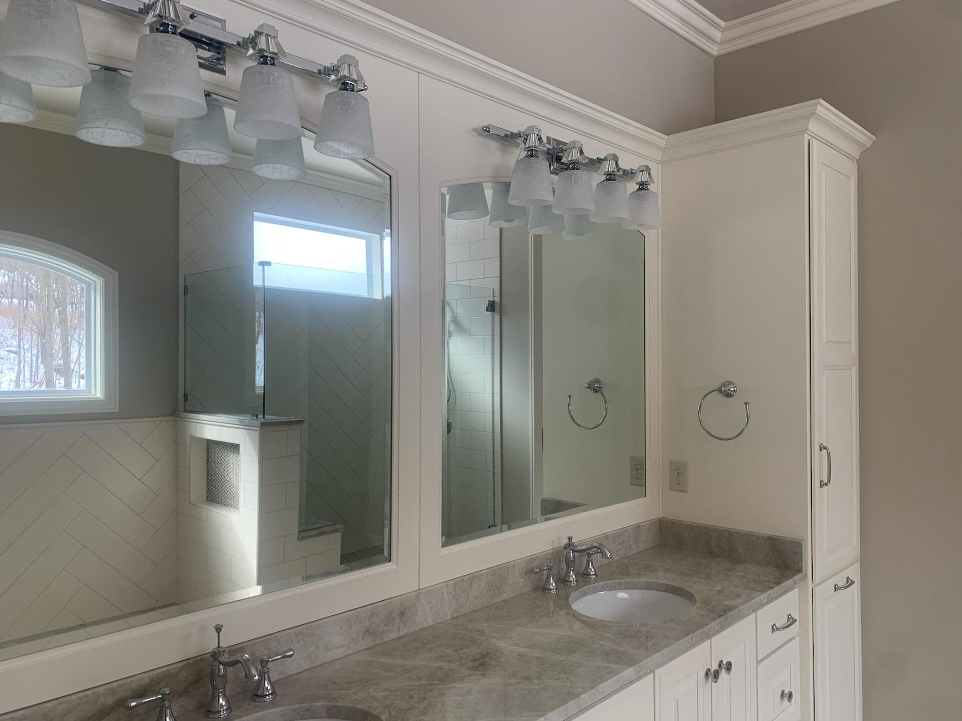 Bathroom with double sinks, mirrors, and a tall cabinet. Light fixtures and a window are visible.