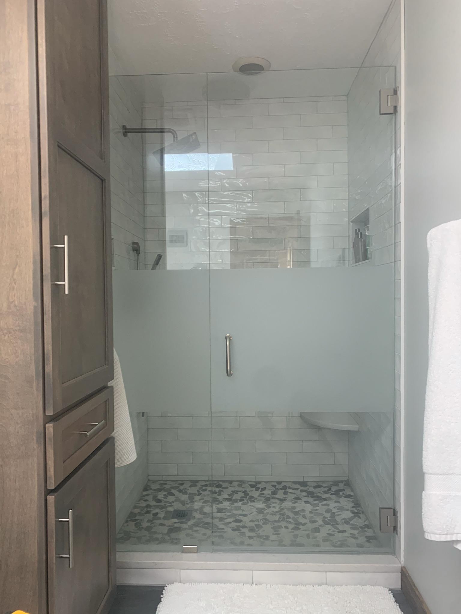 Glass-enclosed shower with frosted glass door and tiled floor; white towel and storage cabinet on either side.