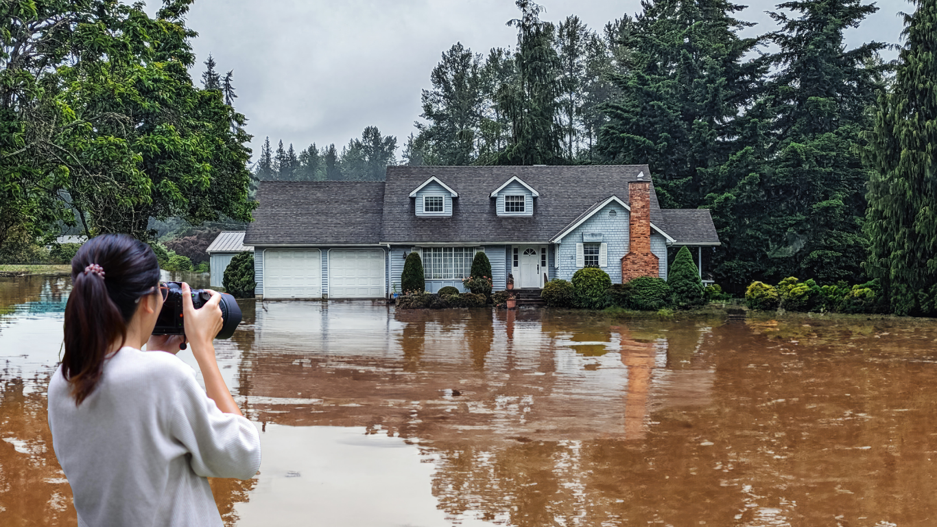 Woman photographs a flooded house. Brown water surrounds the home; it is a gray, overcast day.