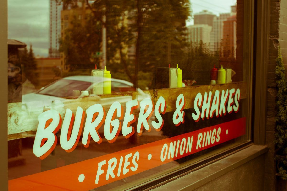Window of a restaurant advertising Burgers & Shakes, with orange accents, condiments visible inside, and city reflected in window.