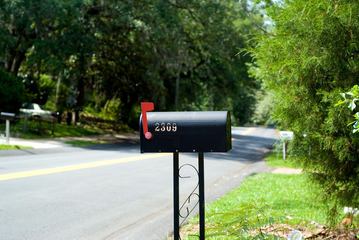 Black mailbox with the flag up by the roadside, with trees and a road in the background.