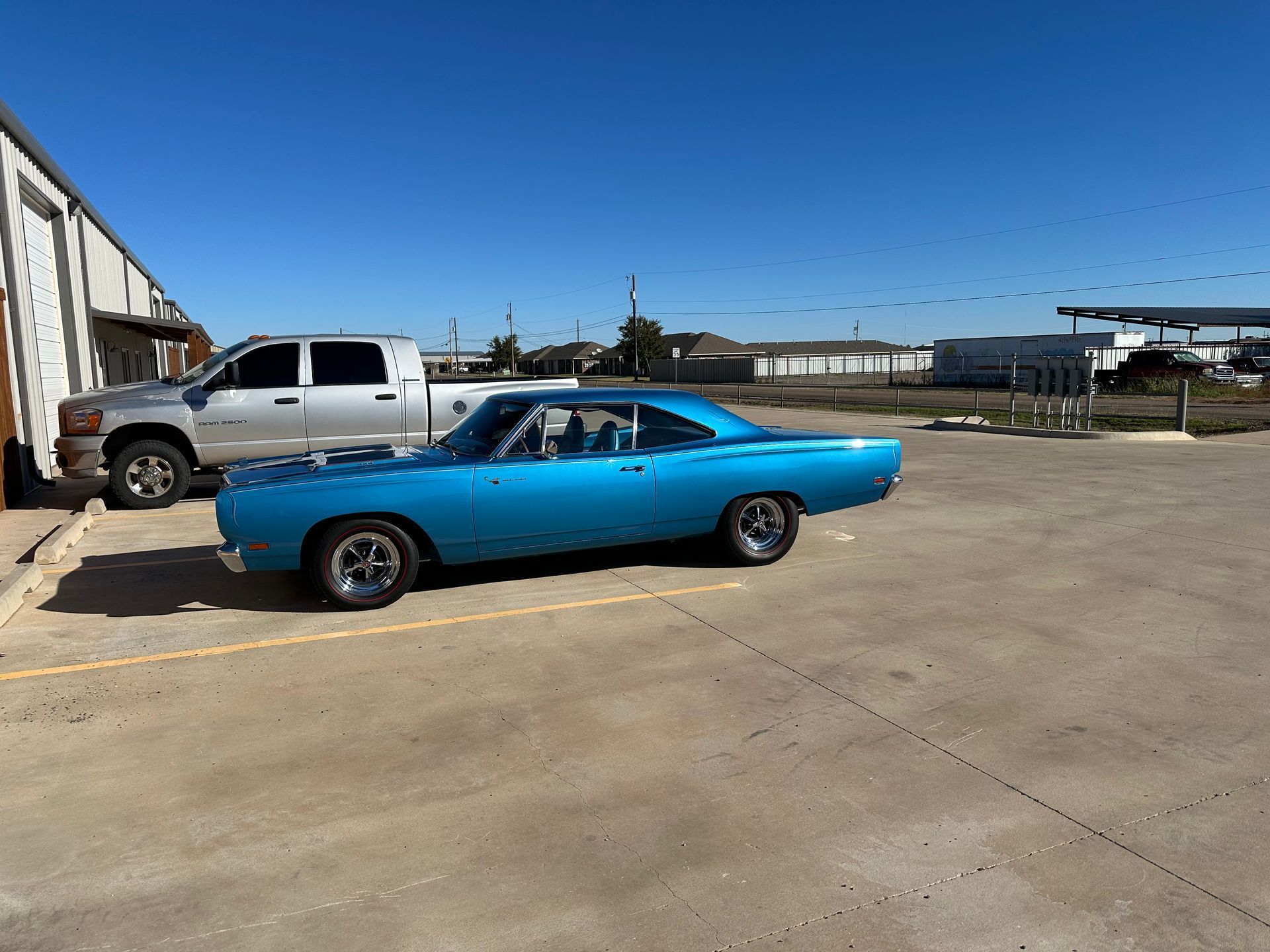A classic turquoise Plymouth Road Runner and silver pickup truck parked outside a building on a sunny day.