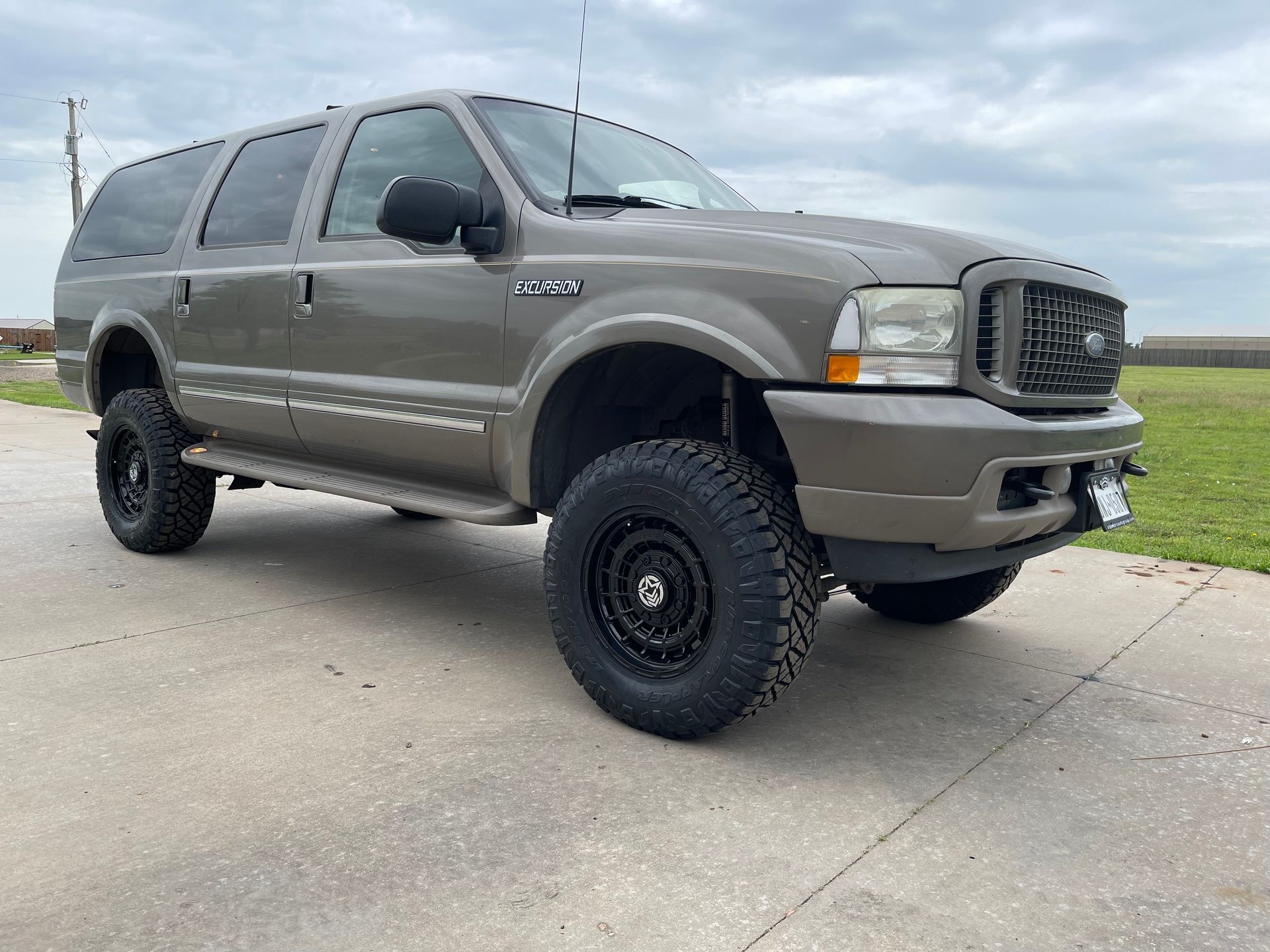 Tan Ford Excursion SUV with black wheels on a concrete surface, cloudy sky.