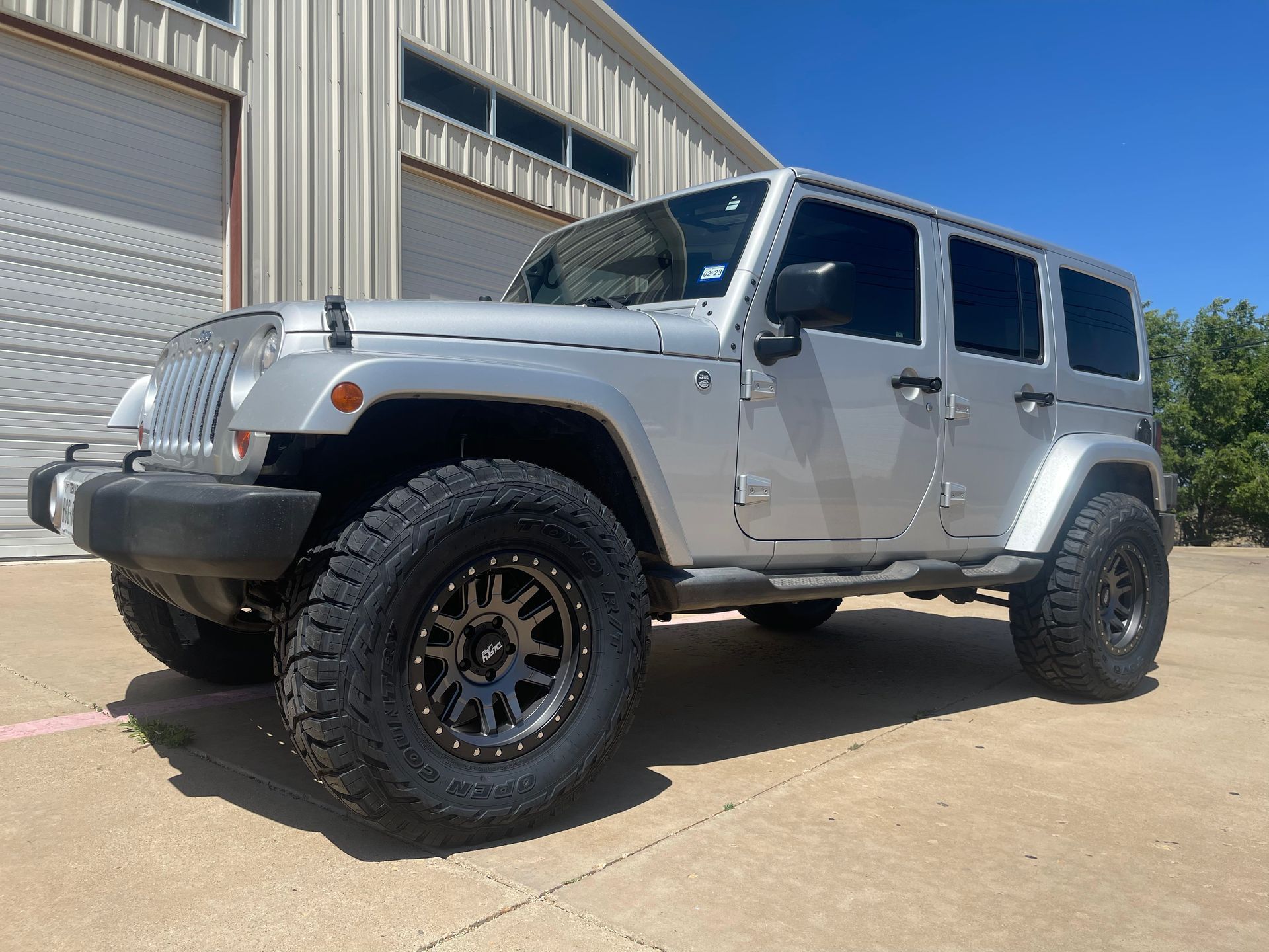 Silver Jeep Wrangler with black wheels, parked in front of a metal building on a sunny day.
